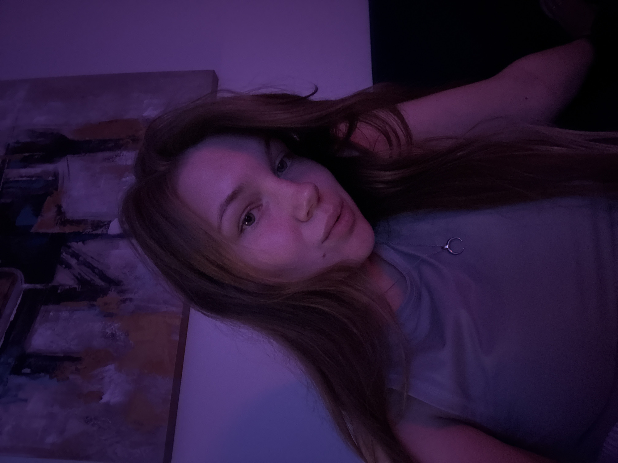 A young woman with light skin and long brown hair is lying down. She is wearing a light-colored t-shirt and a necklace with a small circular pendant. To the left of the woman is a piece of abstract art. The background is dark, with a black area in the upper right corner. The image has a purplish hue, likely due to the lighting in the room. The photo was presumably taken indoors. There is no information that suggests the image was taken in Belgrade, Serbia.