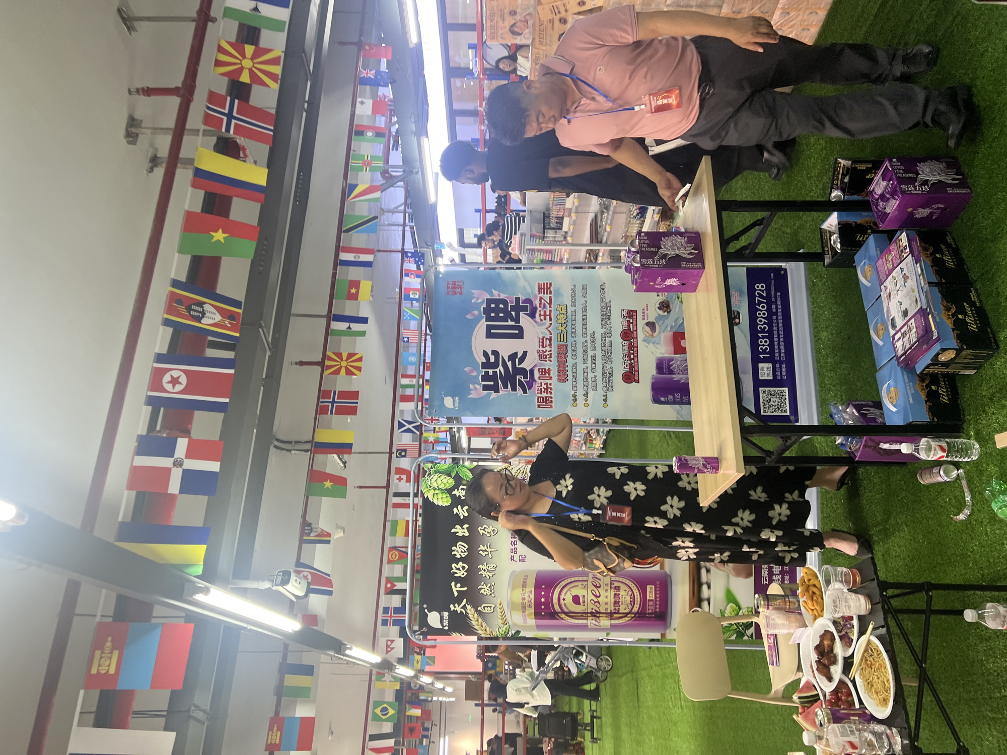 The image depicts an indoor event, possibly a promotional display or market, set up on artificial grass. The background features a ceiling with industrial lighting and a network of pipes. Several strings of national flags are hung across the ceiling, suggesting an international theme.
In the foreground, a woman in a black floral dress stands near a table, adjusting her glasses. On the table are various items including food, drinks, and promotional materials. To the right of the woman, a man in a pink polo shirt and dark pants stands, also near the table, appearing to interact with items on it.
Behind the table, there are large vertical banners with text and graphics, one of which advertises "紫啤" (Purple Beer). Several purple boxes and cans are visible, reinforcing the beverage promotion. Other people are present in the background, some browsing, others working at what appears to be a counter or stall. The overall scene suggests a commercial or exhibition environment. The mention of Liandu District, China, indicates a potential geographic location for this event.
