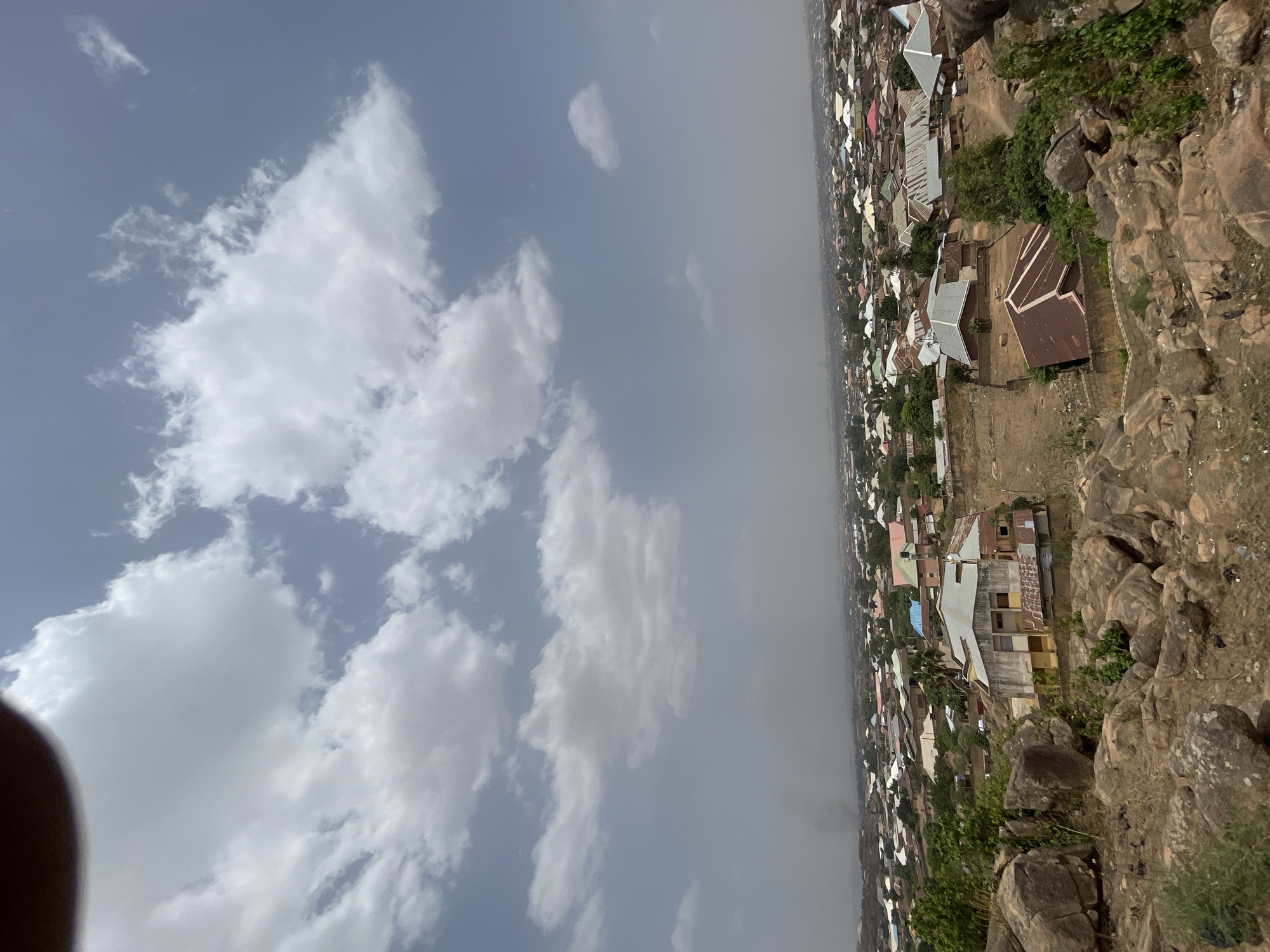 The image shows an elevated view overlooking a residential area in Jos, Nigeria. The sky is partly cloudy. In the foreground, there are rocky formations covered in sparse vegetation. The residential area consists of closely packed buildings with various roof colors, mainly red, brown, and white. The perspective suggests the photo was taken from a higher elevation, possibly a rocky hilltop. The air quality appears somewhat hazy, obscuring distant details. No people are visible.