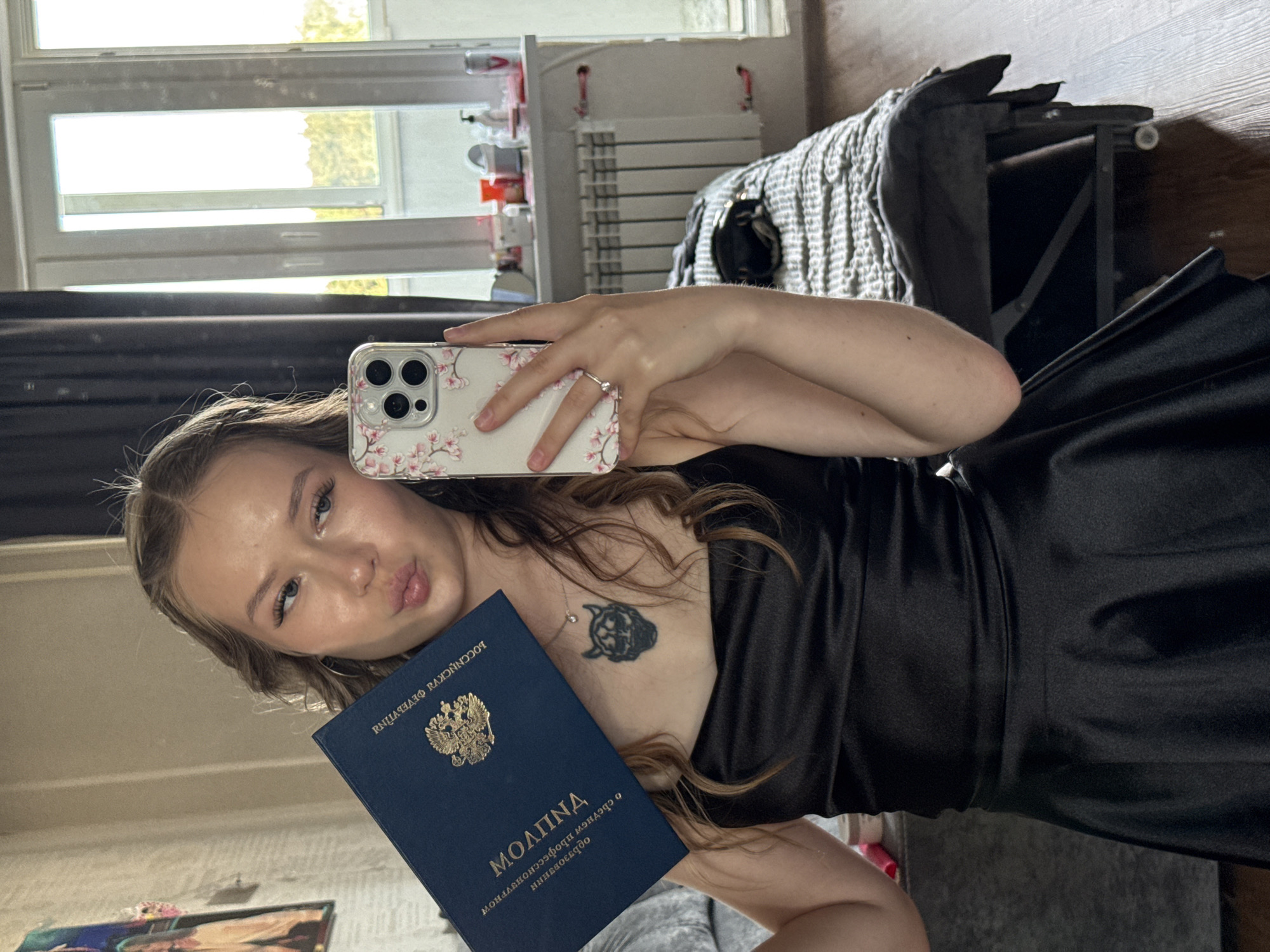 The media file features a young woman taking a selfie in a room. She is holding a dark blue booklet with the Russian Federation's coat of arms and text in Russian visible on it. The booklet appears to be an official document, possibly a diploma or certificate, with Cyrillic text that includes "РОССИЙСКОЙ ФЕДЕРАЦИИ" and "МОЛИТВА". She is wearing a black satin dress and has a tattoo on her chest. Her hair is long and wavy, and she has makeup on. She is puckering her lips, suggesting a playful or affectionate gesture.
The setting appears to be a bedroom, with a bed and a window visible in the background. The window shows sunlight streaming in, suggesting it is daytime. The room is cluttered with items, including what looks like a radiator and shelves with various objects on them. The overall impression is a personal and informal moment captured by the woman. The presence of the official document and the woman's attire suggest she might be celebrating an academic achievement or a significant personal milestone. The selfie is taken with a smartphone.