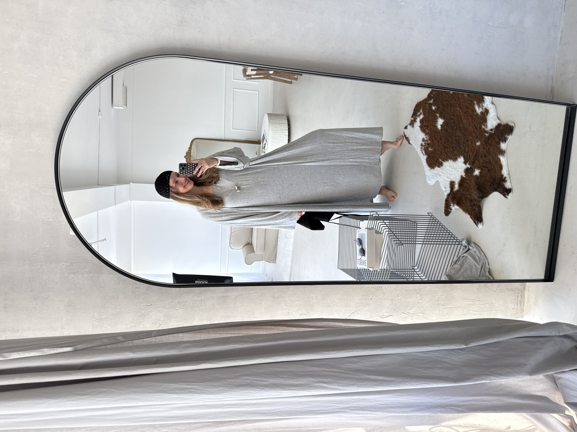 In a bright, minimalist studio space, a young Caucasian woman with long blonde hair and a black beanie takes a mirror selfie. She is wearing a long, loose-fitting grey dress and a necklace with a heart-shaped pendant. She is barefoot and standing on a light-colored floor. To her left, a tall, grey curtain hangs from the ceiling.
Reflected in the large, arched mirror, the room appears spacious and uncluttered. White walls with subtle textured details and baseboards frame the space. In the reflection, a cream-colored sofa with slipcovers is visible in the background, along with a round, white ottoman and a tall, ornate gold mirror. A white rug is partially visible under the sofa.
In the foreground of the reflection, a black metal wire shelving unit holds a few books and a pair of sunglasses. To the right of the shelving unit, a brown and white cowhide rug is spread across the floor. A professional lighting setup is visible in the far right of the reflection, positioned behind the mirror. The overall impression is one of a modern, airy, and well-lit interior. The presence of the lighting equipment suggests the space is likely a photography studio or a similar creative environment. The location is not specified as being in Izhevsk, Russia.