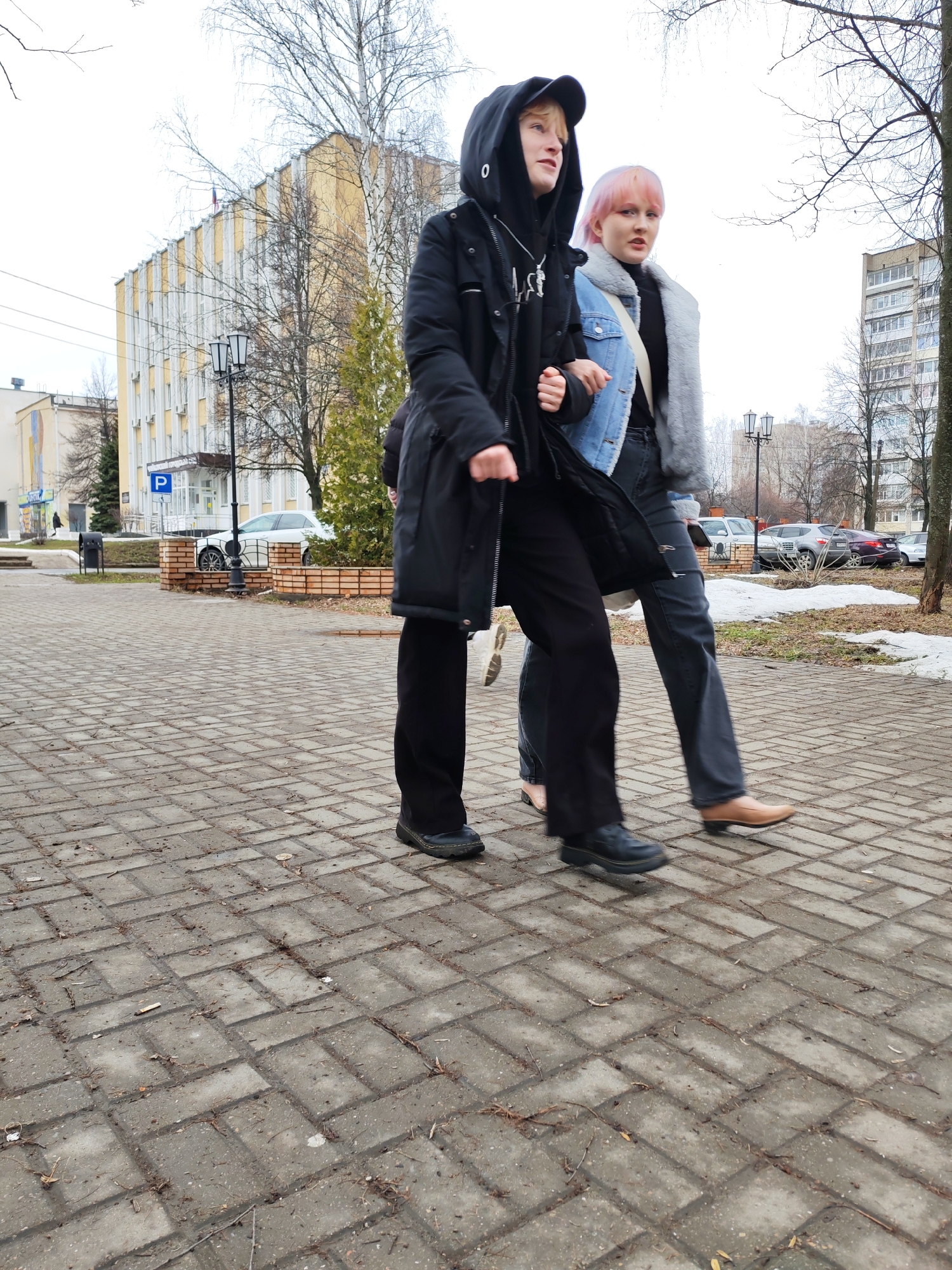 The image shows two individuals walking side-by-side on a paved sidewalk. The first individual, on the left, is wearing a black hooded jacket and dark-colored pants and shoes. The second individual, on the right, has pink hair, wears a denim jacket with a grey faux fur collar, dark jeans, and tan shoes. They appear to be walking in the same direction.
The location is a paved area with a building in the background. A "P" parking sign is visible, as well as a building with classical architectural features. Cars are parked in the background. Trees, bare of leaves, are also visible. The presence of snow patches suggests it is late winter or early spring.
The presence of the "P" sign and the architecture of the building are consistent with a location in the city of Chaikovsky, Russia.