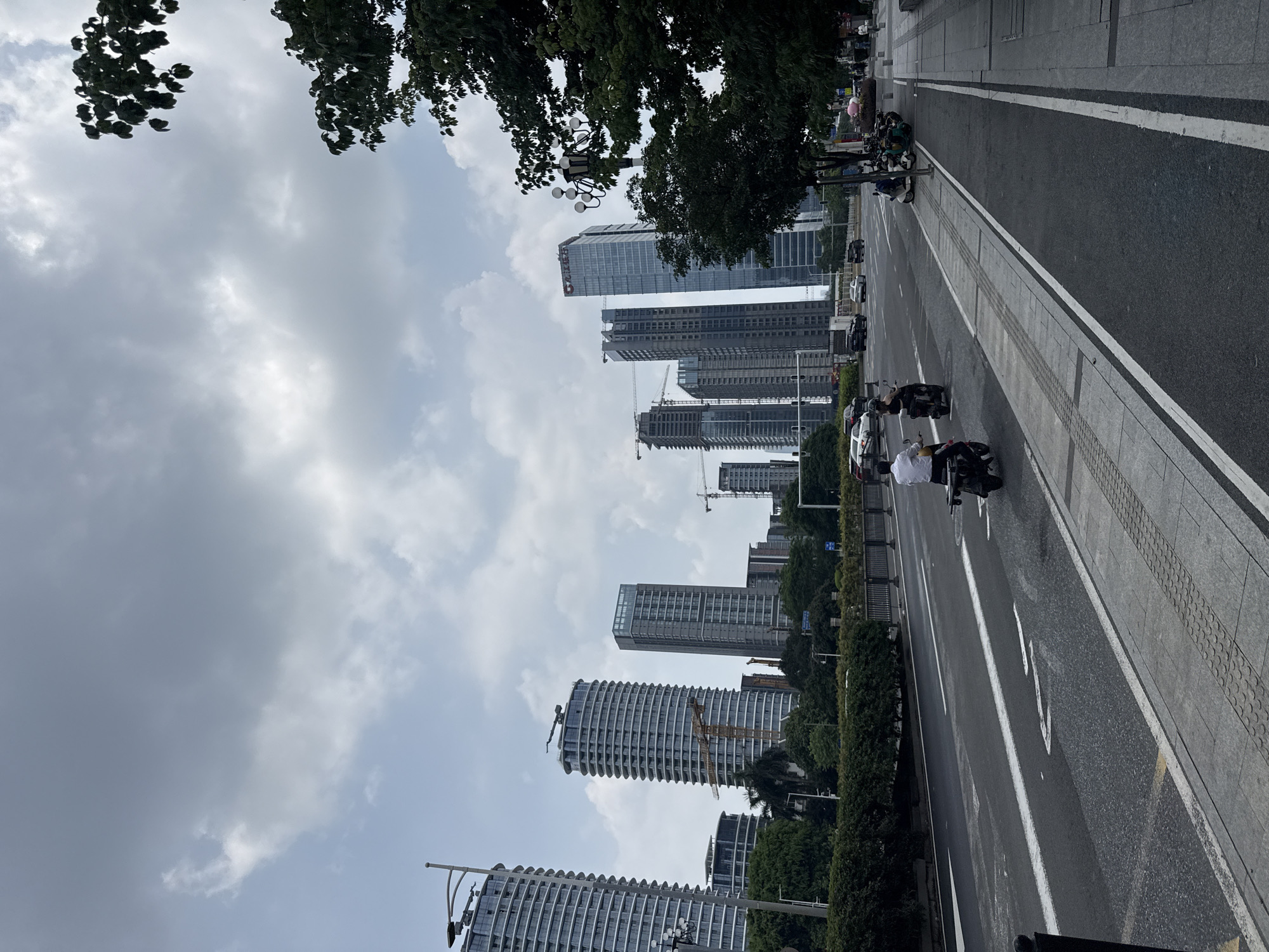 The image captures an upright, bustling urban street scene dominated by modern high-rise architecture under an overcast sky.
**Subjects:**
* **People:** Several individuals are visible on the road and sidewalk, primarily riding or standing by motorcycles and scooters. One person in a white shirt on a dark scooter is clearly visible in the mid-ground.
* **Objects:** The skyline is filled with numerous tall, contemporary buildings, many featuring glass facades. Several of these structures are under active construction, indicated by multiple yellow tower cranes extending above them. Some buildings exhibit distinctive designs, such as a prominent cylindrical building with repetitive, stacked balcony elements. The foreground features a multi-lane asphalt road with clear white lane markings, including a partial directional arrow. Adjacent to the road is a wide sidewalk with tactile paving. A significant number of motorcycles and scooters are parked along the sidewalk or are in motion on the road. A few cars are also visible further down the street. Green foliage from trees frames the top of the image and lines the street, while dense bushes form a hedge along a central reservation. A multi-globed street lamp is present on the left side of the road.
* **Scene:** A vibrant, developed city environment with ongoing urban expansion.
* **Setting:** An urban landscape.
**Activity or event taking place:**
The scene depicts daily urban life, with vehicles and people traversing the city streets. The presence of construction cranes highlights ongoing development and growth within the city.
**Notable details:**
* **Time of day:** It is daytime, under a bright but heavily overcast sky with varying shades of gray clouds.
* **Weather:** Cloudy conditions prevail, but there are no signs of precipitation.
* **Location cues:** The modern high-rise buildings and the prevalence of two-wheeled vehicles suggest an Asian metropolis, consistent with the Tianhe District, China context.
**Visible text:**
A red logo or text, likely Chinese characters, is visible on the upper part of a prominent glass-faced high-rise building in the background. The specific characters are difficult to discern clearly but appear to be a corporate or brand name.
