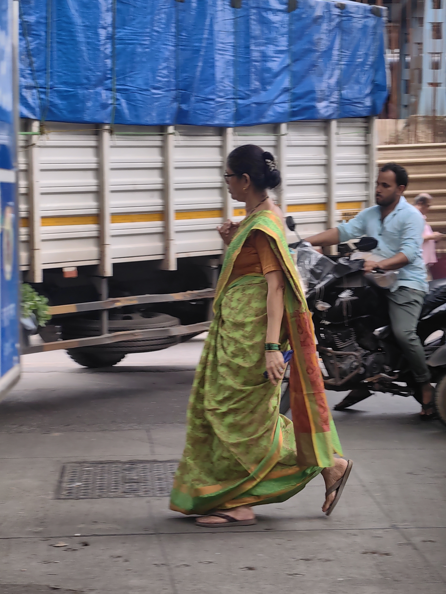 In Mira Bhayandar, India, a woman in a green and orange sari walks on a city street. She is wearing glasses and has her hair pulled back in a bun. She is also wearing flip-flops. Behind her, a man rides a motorcycle. He is wearing a light blue shirt and green pants. A large truck is parked behind the motorcycle. The truck has a blue tarp covering its cargo. The street is paved, and there is a grate in the pavement. The overall scene depicts a typical day on a busy street in India.