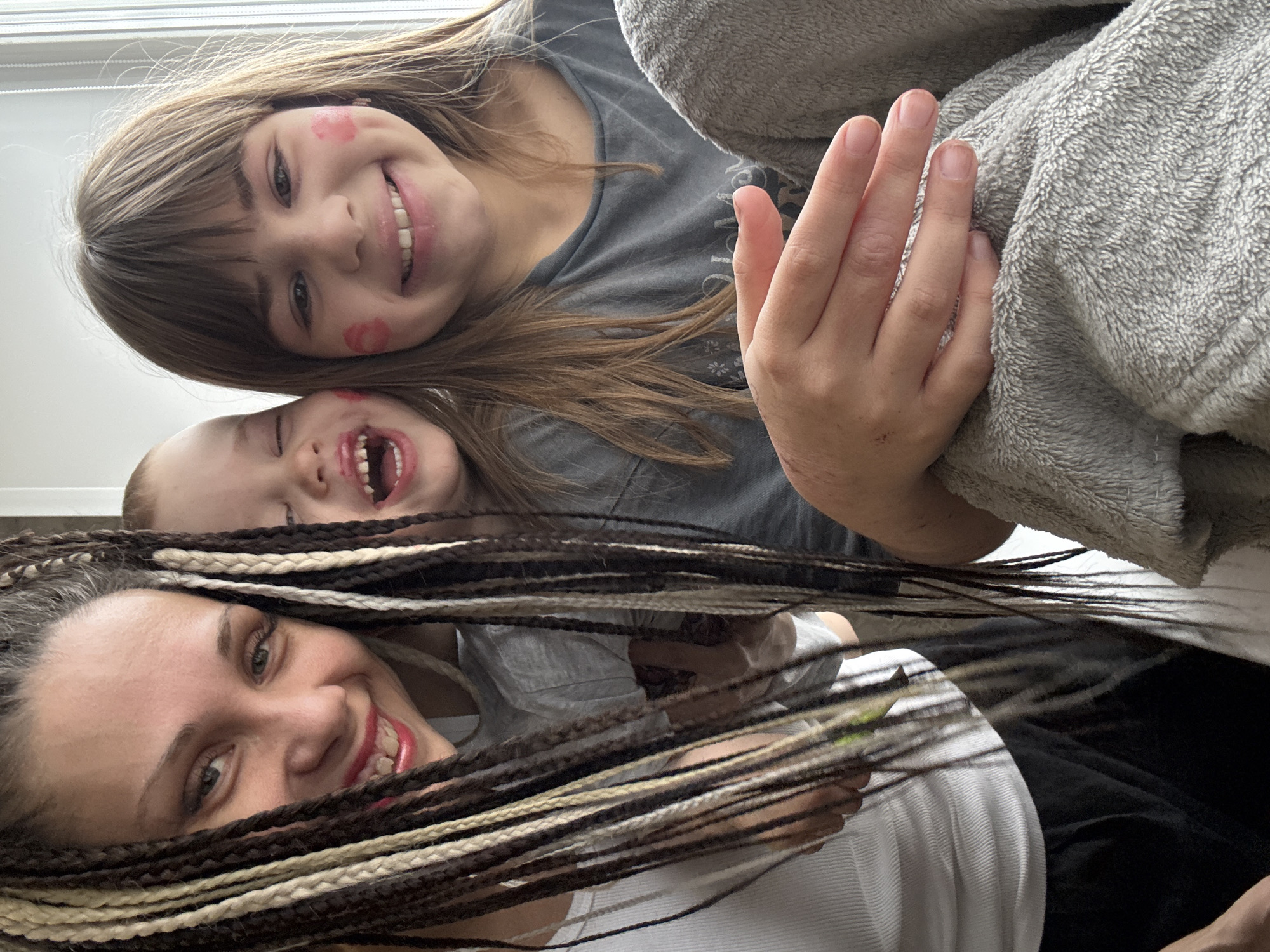 A vertically oriented, close-up image captures a cheerful moment among three individuals, likely indoors. In the foreground, a woman looks upwards and smiles broadly. She has long, intricate braids (dreadlocks or box braids) in brown and blonde tones, framing her face, and is wearing a white ribbed top.
Behind her, an older child, likely a girl, also smiles widely, laughing with an open mouth. She has shoulder-length brown hair with bangs and sports playful red lipstick kiss marks on her cheeks. She is dressed in a grey t-shirt, and her hand, with natural nails, is visible resting on a soft, grey, fuzzy blanket or robe.
Partially visible behind the first child is a second, smaller child, also captured mid-laugh with an open mouth, suggesting shared amusement and joy. They also appear to have red marks on their face. The image is taken from a low angle, emphasizing the close interaction and happiness among the group. No visible text is present. The setting appears to be a home in Rusanovo, Russia.