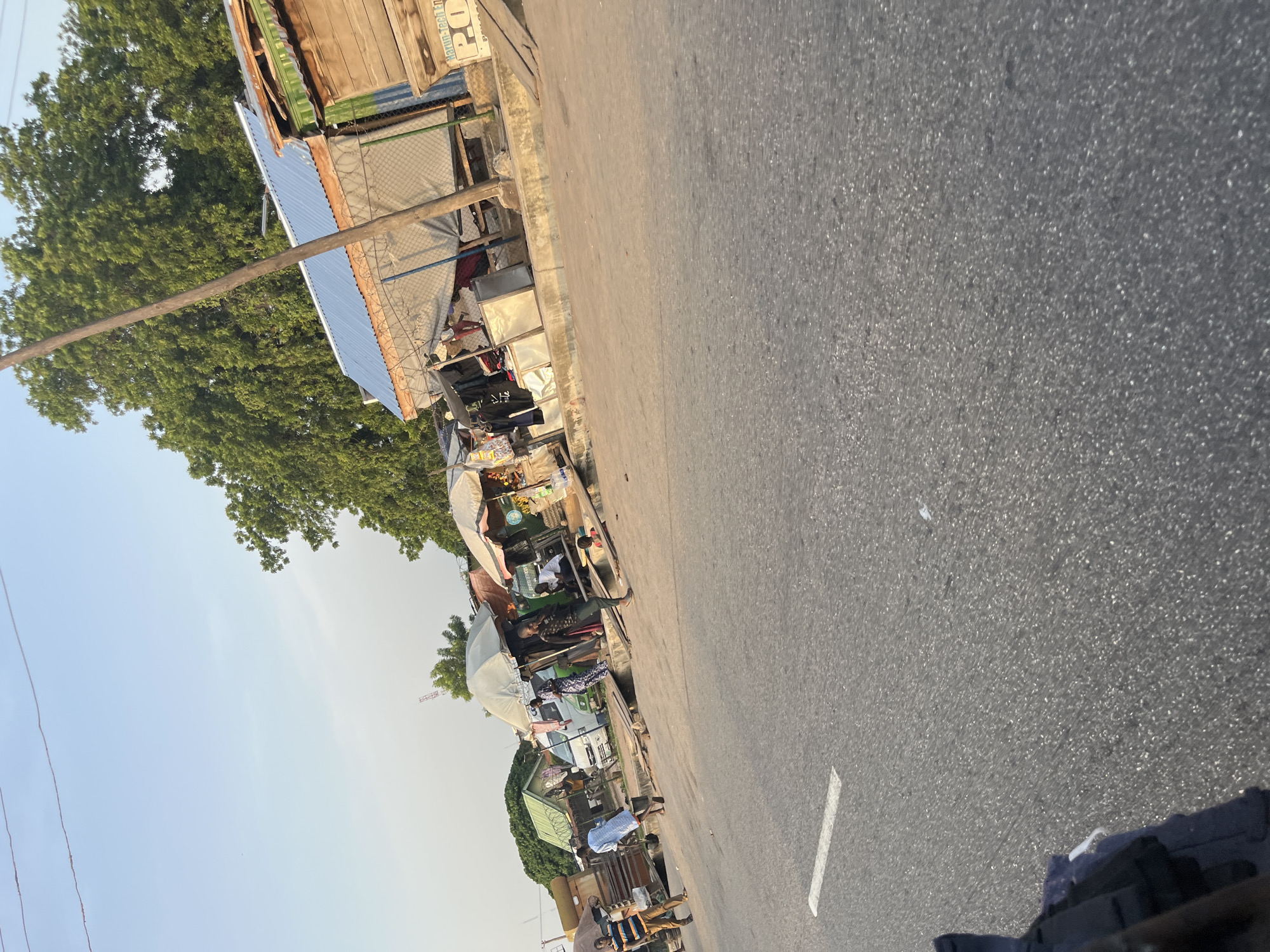 A street scene with several stalls and people is captured from a moving vehicle. The road is paved and has a white lane marking. On the right side of the road, there are several market stalls with various goods displayed, including clothing and what appears to be food items. Some stalls have awnings for shade. Several individuals are seen standing around the stalls, some interacting with each other and others seemingly browsing. A white van is parked behind some of the stalls. Tall green trees are visible in the background. A wooden utility pole stands near the stalls. The sky is clear and blue. The lighting suggests it is daytime, possibly late afternoon due to the warm tones.