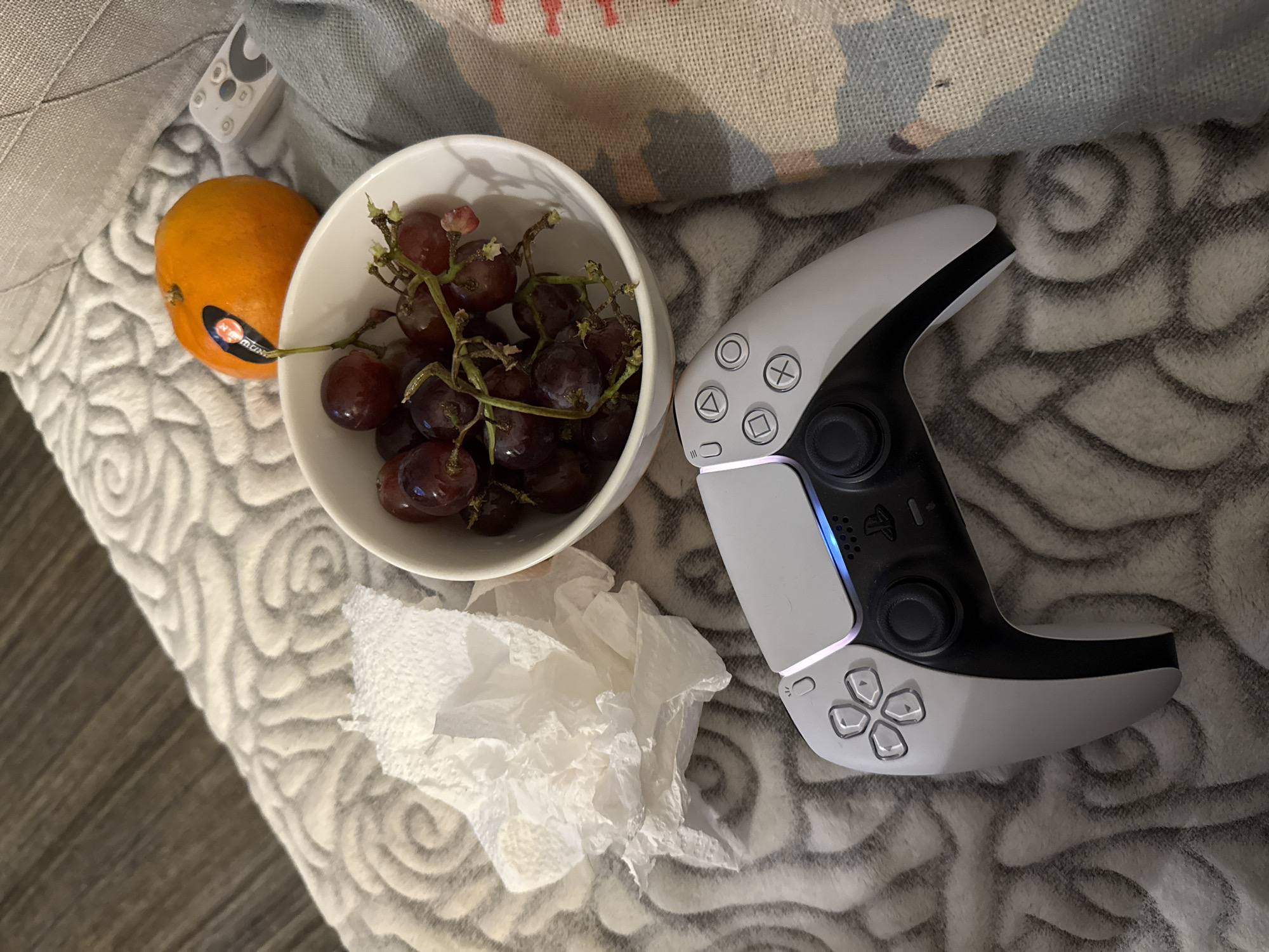 The image shows a still life scene on what appears to be a textured blanket or upholstered surface. A white ceramic bowl is filled with dark red grapes, positioned next to an orange with a sticker. A crumpled white paper towel lies below the bowl and grapes. To the right of the grapes and paper towel is a PlayStation 5 DualSense controller. The lighting appears to be indoor, potentially from artificial sources. There is no evidence to suggest that the image was taken in Petah Tikva, Israel.