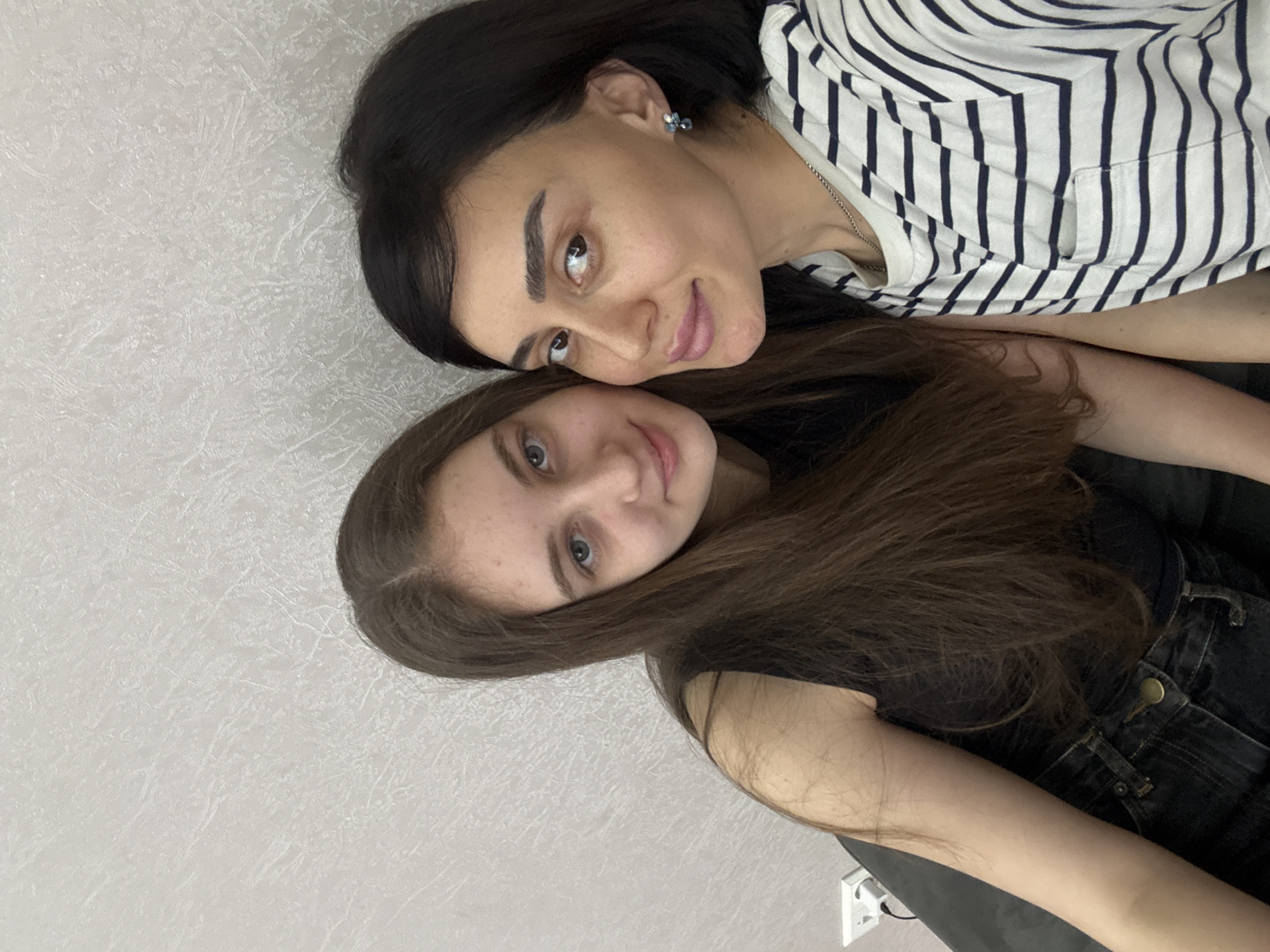Two females are closely positioned in the frame. The upper female has dark hair and is wearing a striped shirt. The lower female has brown hair and appears to be wearing a black outfit. The setting is indoors against a textured white wall. The location of the city of Tbilisi, Georgia is not identifiable.