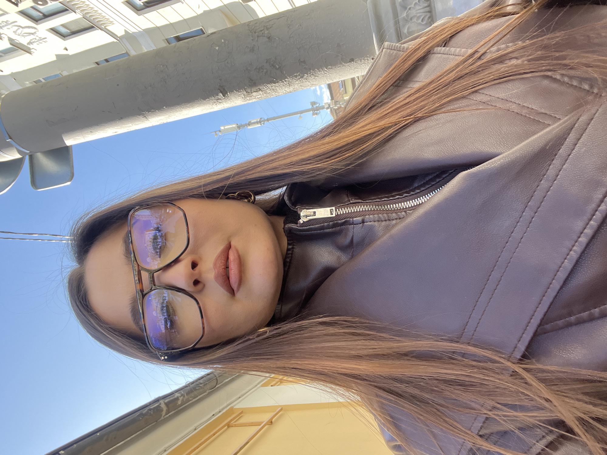 A woman is photographed from a low angle outdoors in Moscow, Russia. The woman wears glasses and a brown leather jacket. Buildings and blue sky are visible in the background.