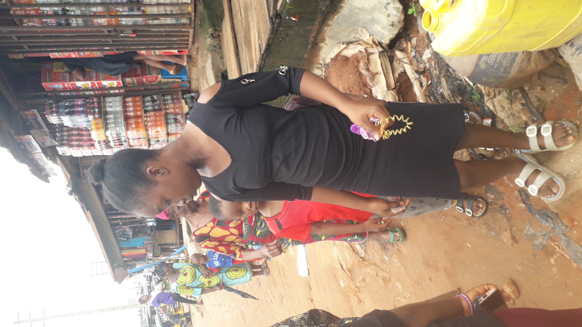 The image shows a scene in Nkpor, Nigeria. In the foreground is a woman wearing a black dress and white sandals. She is holding a gold bracelet and a purple item. Behind her, a woman in a patterned dress stands next to a child wearing a red shirt. In the background, there is a shop filled with stacks of bottled drinks. The ground appears to be dirt or clay. A yellow container sits near the woman's feet. Other people are seen in the background, engaging in various activities.