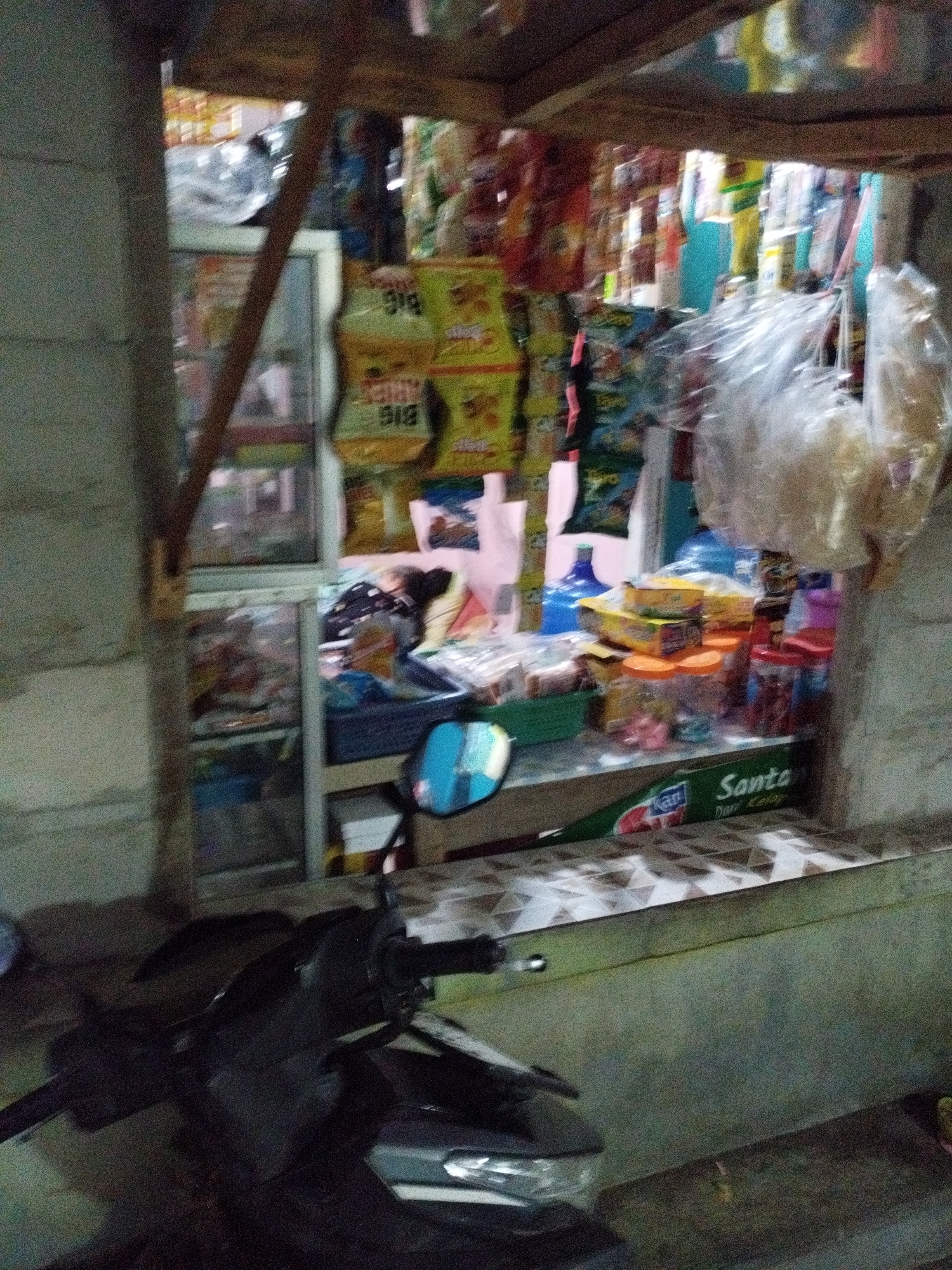 The image shows a small, open-air convenience store, likely located in Kelurahan Cilacap, Kecamatan Cilacap, Indonesia. The store appears to be built with cinder blocks and has a wooden overhead structure. It is stocked with various packaged snacks, drinks in bottles, and other assorted goods, many of which are displayed on shelves or hanging. In the foreground, the handlebars and front of a dark-colored motorcycle are visible. A person, possibly a store attendant or customer, is partially visible behind the counter, lying down. The overall scene suggests a typical local shop operating in a residential or commercial area. The lighting indicates it may be evening or the store is dimly lit.