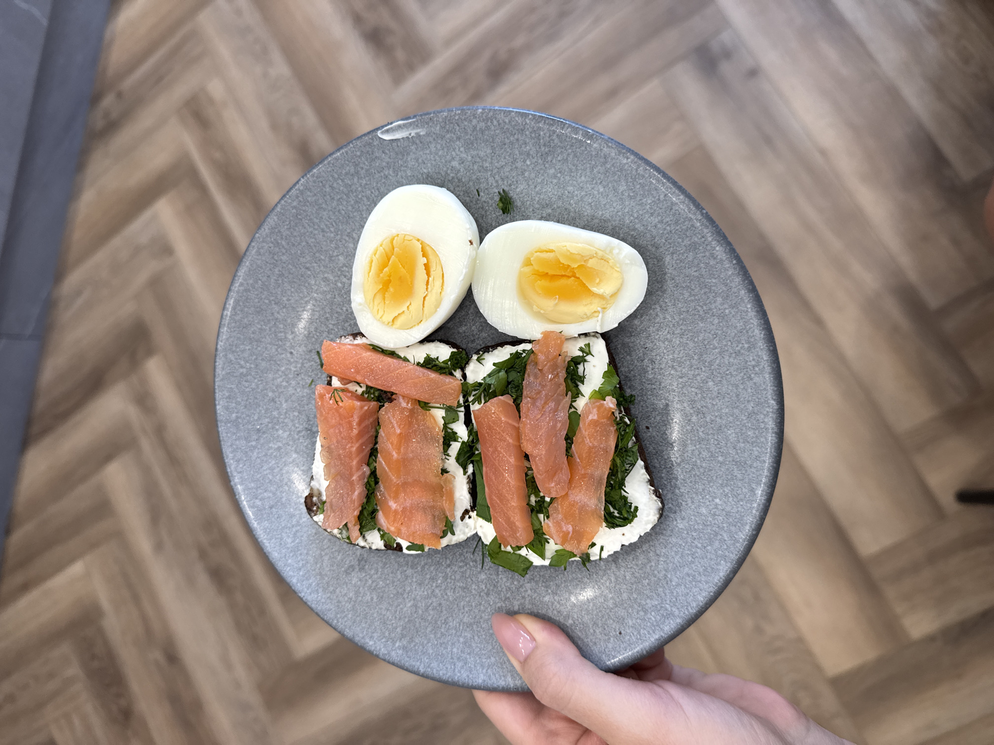 A person is holding a grey plate of food consisting of two pieces of toast topped with a white spread (possibly cream cheese), green herbs, and slices of salmon. Beside the toast are two halves of a hard-boiled egg. The background shows a wooden floor with a herringbone pattern. Location: Unknown, Unknown.