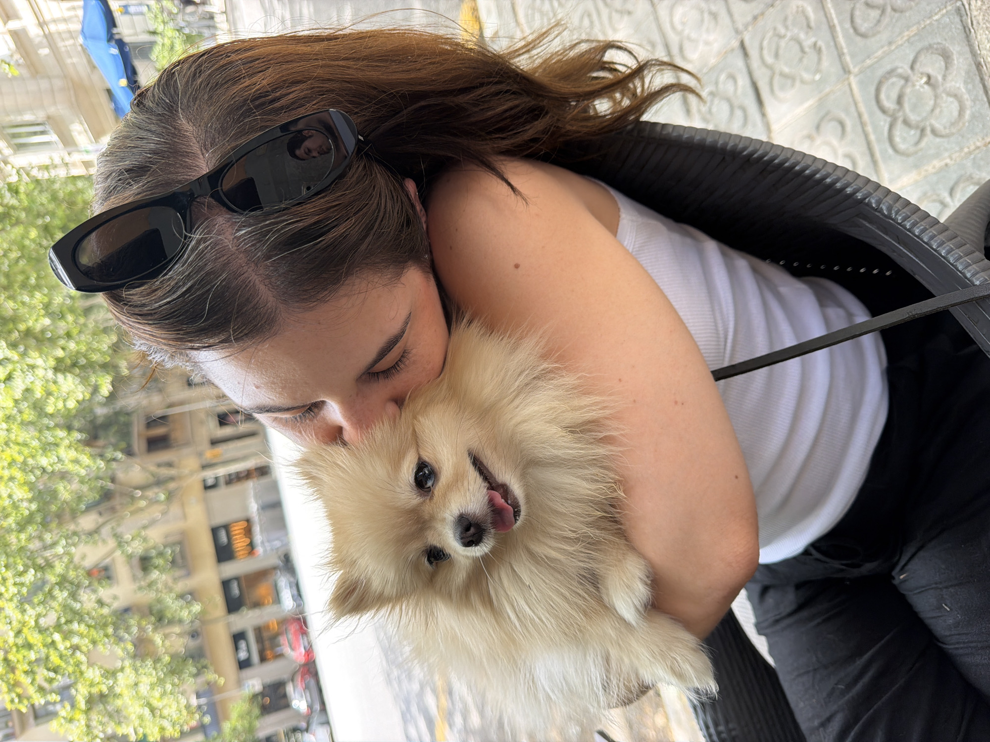 A young woman with long brown hair, wearing a white tank top and black pants, is holding a light brown Pomeranian dog on her shoulder, kissing its head. She is wearing black sunglasses perched on top of her head. The background shows a city street with buildings, trees with green foliage, parked cars, and a blue umbrella. The scene appears to be outdoors on a sunny day. The floor tiles are visible in the foreground. The architecture and streetscape suggest a location in Barcelona, Spain, possibly the Eixample district due to the characteristic pavement tiles and building style.