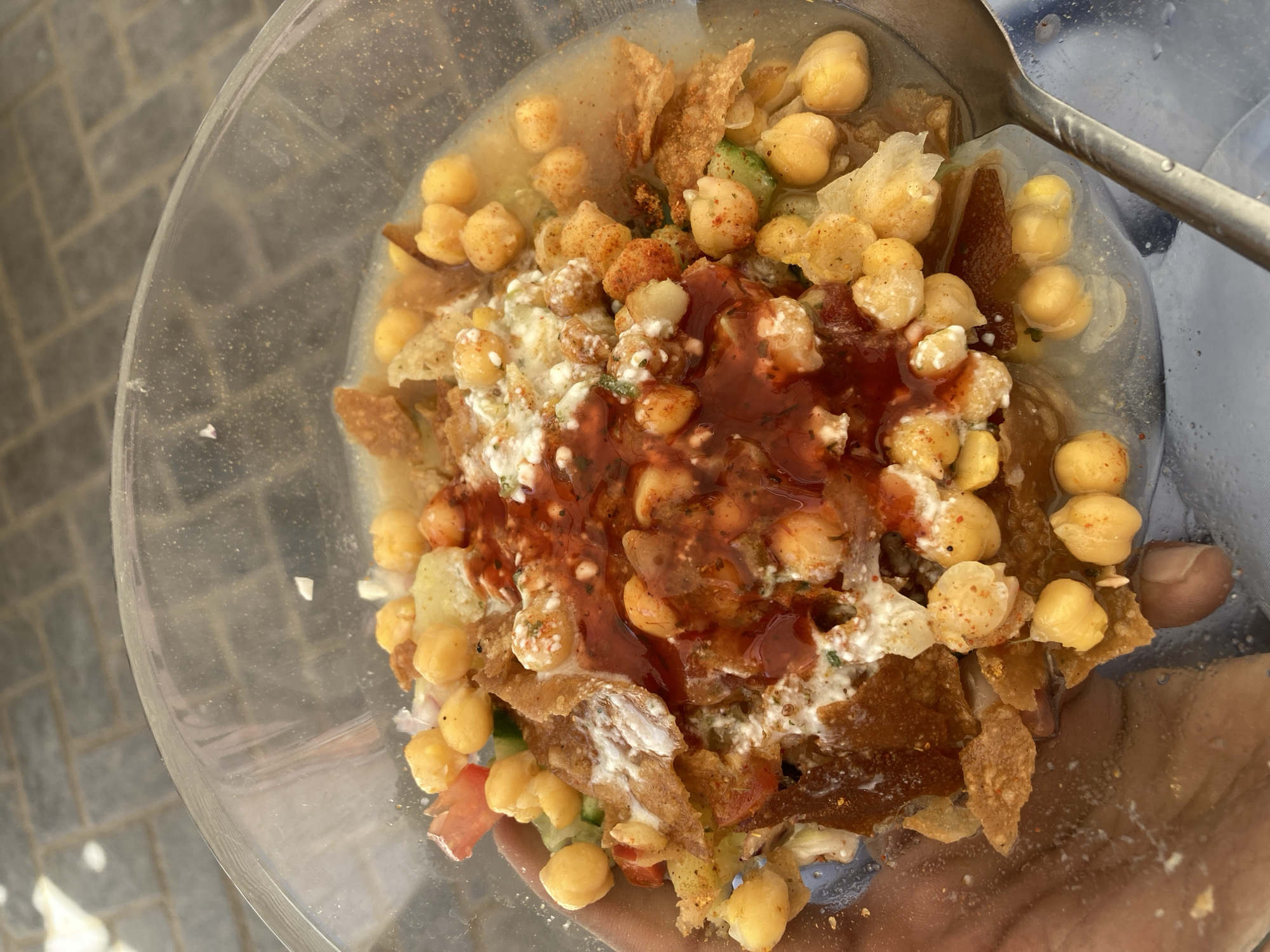 A bowl of chana chaat, a Pakistani street food, is held in someone's hand. The chana chaat is composed of chickpeas, crispy fried dough wafers, potatoes, yogurt, chutney, and spices. The bowl appears to be clear plastic. A metal spoon rests inside the bowl. The location is Quetta City Tehsil, Pakistan. The background is a patterned surface, possibly tile.