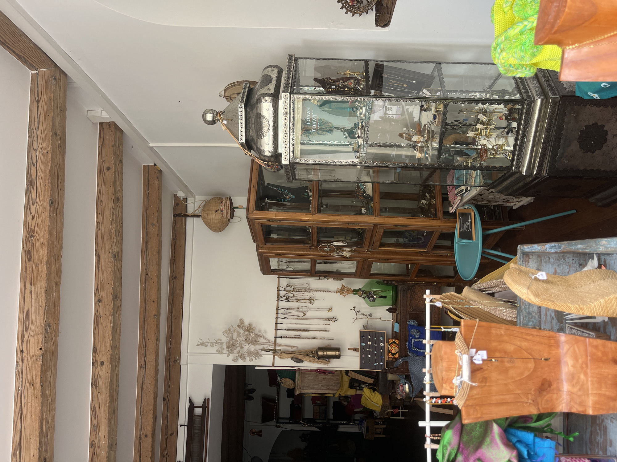 This is an interior shot of a retail store located in Vevey, Switzerland, displaying various jewelry and accessories. The store features rustic wooden ceiling beams and white walls. To the left, a wooden display rack holds necklaces and bracelets. In the center, a tall, glass-fronted wooden cabinet showcases more jewelry, and to its right, a decorative silver lantern-style display case is filled with jewelry. Further to the right, a green glass bottle and other decorative items are visible. In the foreground, wooden display stands hold more jewelry and colorful textiles. The overall atmosphere suggests a boutique specializing in artisanal or ethnic-themed goods. There are no people visible in the image.