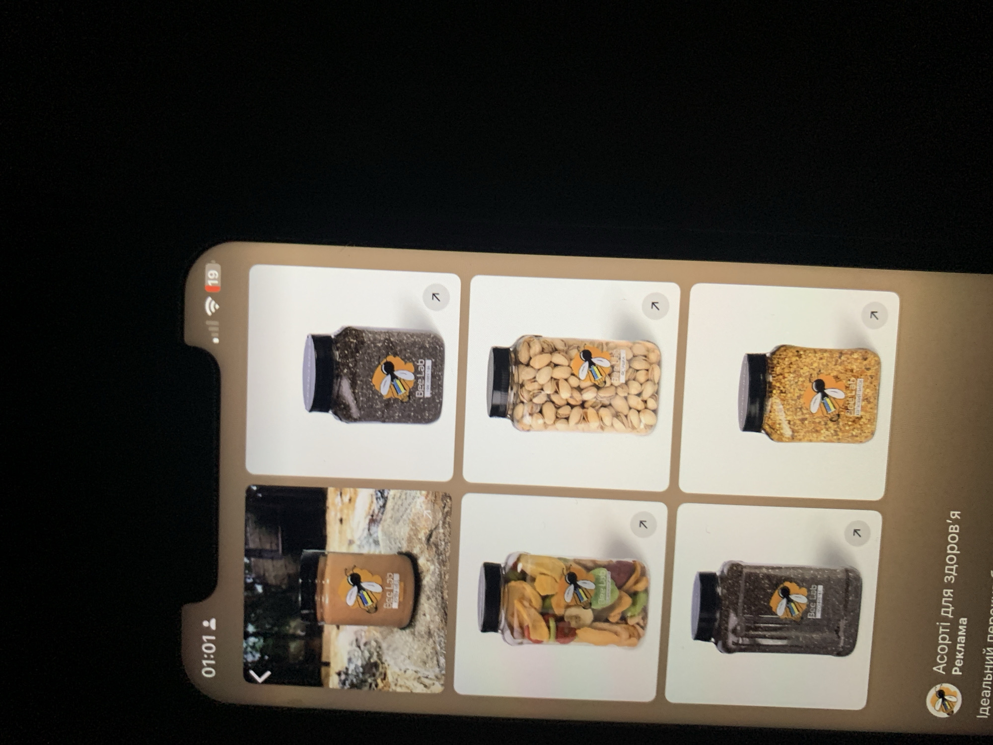 The image shows a mobile phone screen displaying a social media feed or online store. The screen is showing a grid of six product images, each featuring a clear plastic jar with a black lid. The jars contain different food items: the top left jar has a creamy substance, likely peanut butter; the top right jar contains dark seeds, possibly chia seeds; the middle left jar is filled with dried fruit pieces; the middle right jar contains shelled pistachios; the bottom left jar appears to contain more dark seeds; and the bottom right jar is filled with small, granular items, possibly bee pollen or granulated sugar. Each jar has a label with a bee logo and text that appears to be "Bee Lab". The bottom of the screen shows an advertisement with the text "Асорті для здоров'я" (Assortment for health) and "Реклама" (Advertisement). The time displayed at the top of the phone screen is 01:01. There is no information in the image to suggest this scene is located in Zhmerynka, Ukraine.