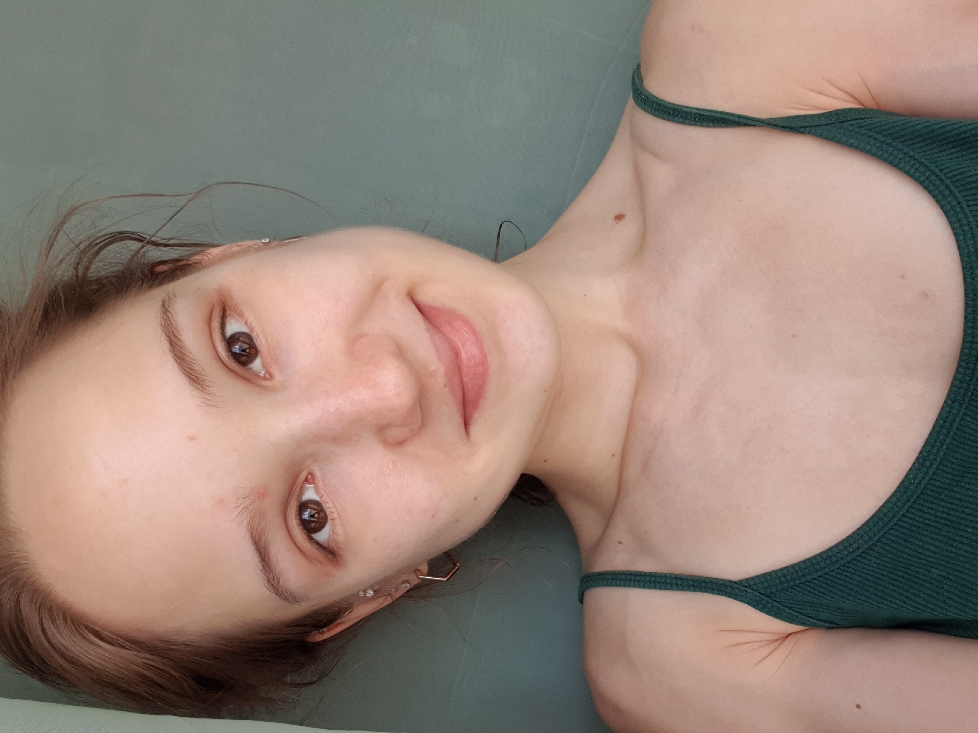 A young woman with light skin, brown eyes, and brown hair is lying on a green surface, possibly a bed or couch. She is wearing a dark green tank top with thin straps. She is looking at the camera with a slight smile. She appears to be wearing earrings. There is no evidence indicating this photo was taken in Khimki, Russia.