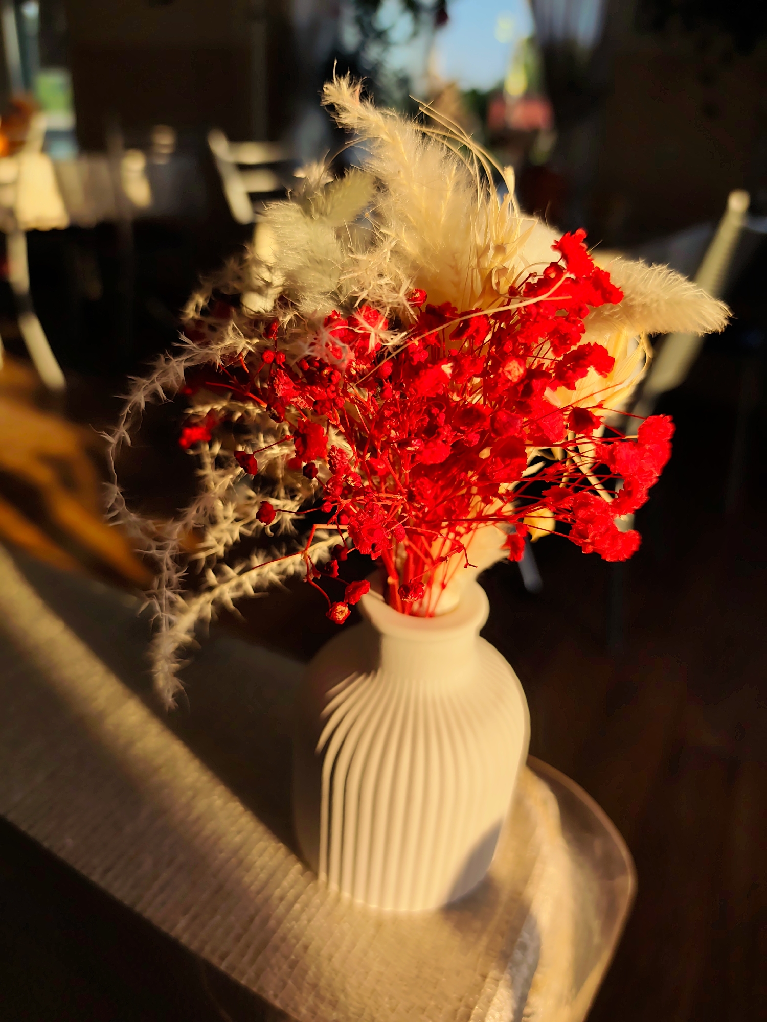 A low-angle shot captures a vase containing dried flowers. The vase is white and features vertical ribbing around its bulbous form. Within the vase, there is a bouquet of dried plants, featuring wispy, cream-colored feathery plumes, delicate white spiky stems, and a cluster of vibrant red dried flowers. The arrangement sits atop a surface covered with a beige, textured fabric, which is illuminated by a strong, warm light from the left side of the frame, casting a long shadow to the right. The background is blurred, showing hints of chairs and tables suggesting an indoor setting. The scene is bathed in a golden light, indicating either sunrise or sunset. There is no indication the scene takes place in Нижний Пальник, Russia.