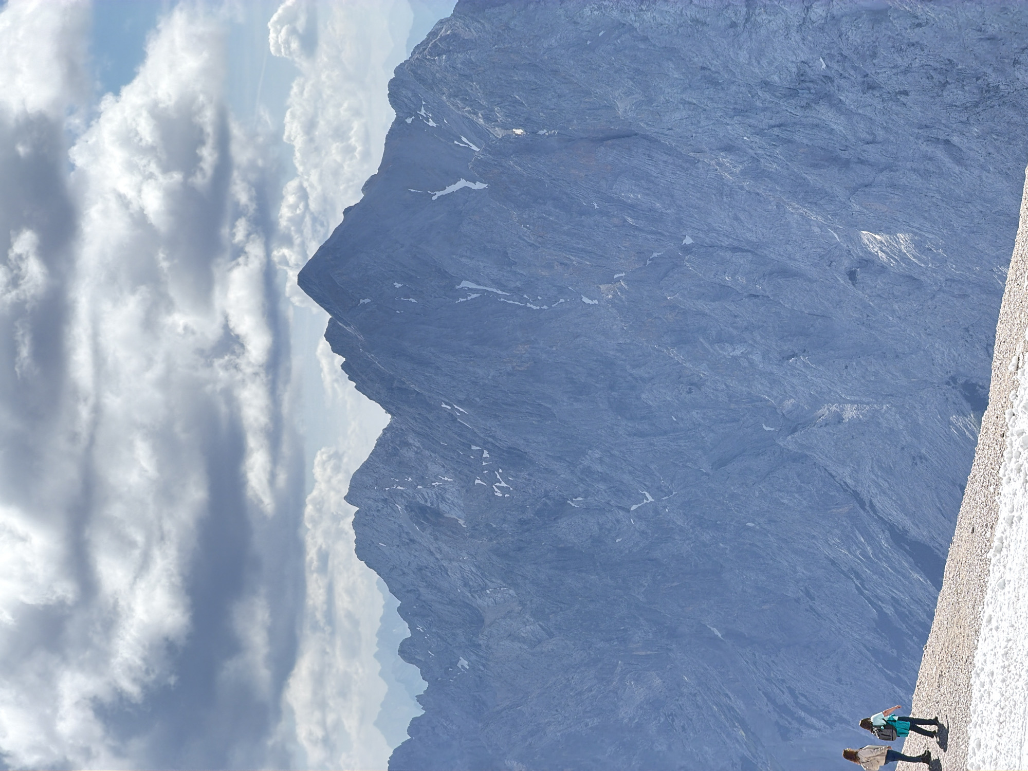 Two women stand on a rocky, snow-dusted mountain slope, facing away from the camera. They are looking towards a vast, blue, rocky mountain range that dominates the background. The mountains are jagged and steep, with patches of snow clinging to their surfaces. The sky above is filled with dramatic, grey and white clouds, with patches of blue sky visible between them. This scene is likely the Zugspitze, Germany's highest peak, located near Garmisch-Partenkirchen.