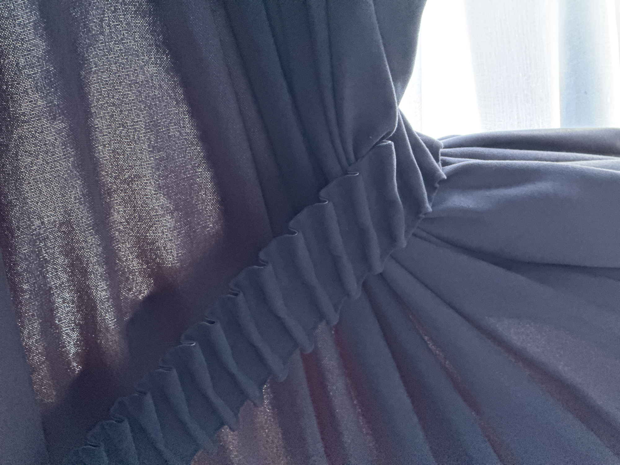 This is a close-up, eye-level, indoor photograph of dark blue curtains. The fabric of the curtains is gathered in the center with a ruffled band. To the left, the curtains are pulled back, revealing a bright white light that appears to be a window with sheer blinds. The background fabric has a subtle shimmer, suggesting a textured or metallic thread woven into it. The image appears to be taken in a domestic setting, likely a home or hotel room, not in a public space in Doma 1096 Km, Russia. There are no people or discernible actions captured in the frame.