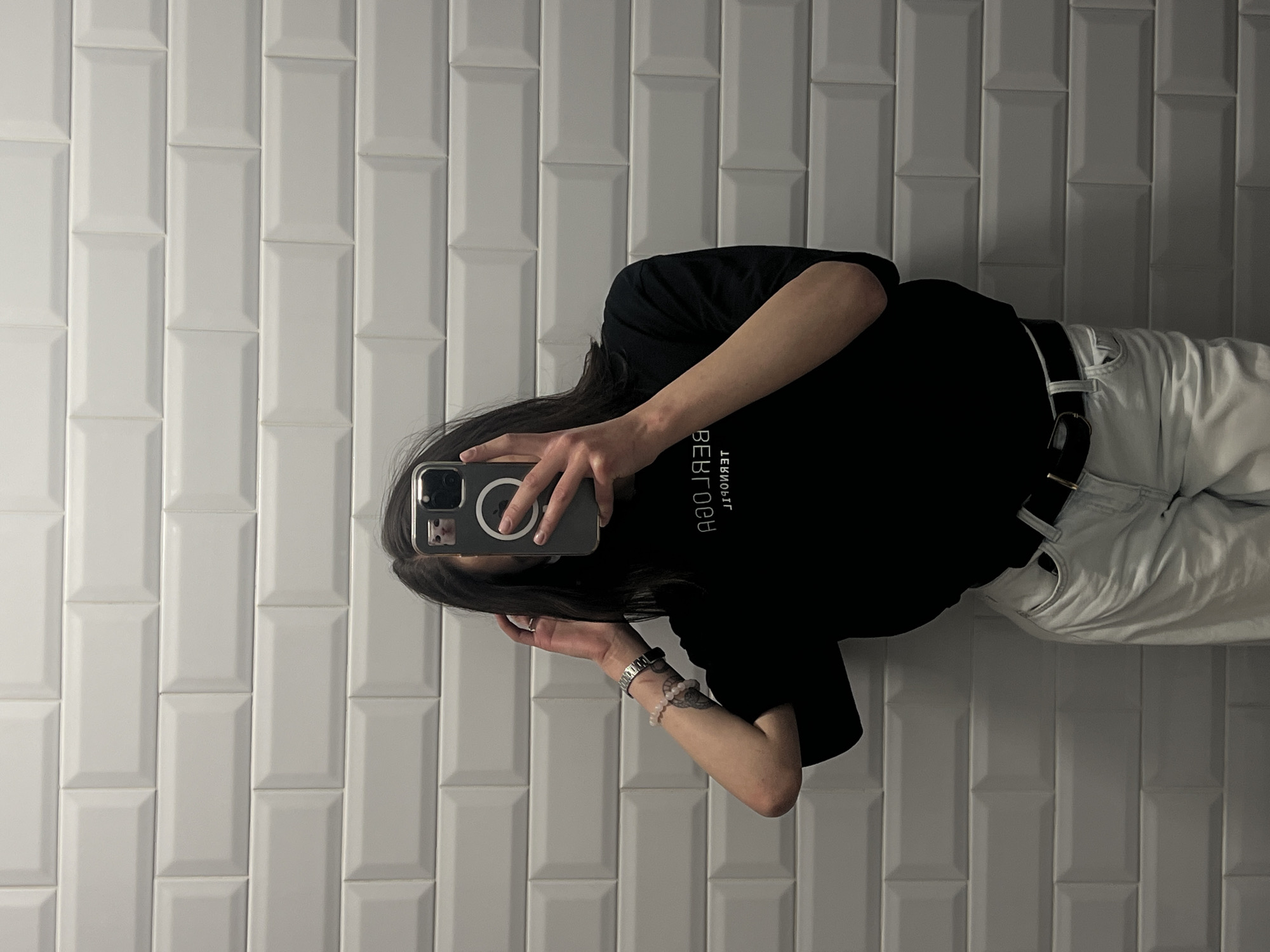 A person is taking a mirror selfie in front of a white tiled wall. The person is wearing a black t-shirt with white text, light-colored jeans, and a black belt. They have dark hair, are wearing a watch on their left wrist, and have bracelets on their left arm. A smartphone with a small cat sticker is visible in their hand, being used to take the photo. The environment appears to be an indoor space with a tiled wall that resembles a bathroom or kitchen. The text on the t-shirt is reversed due to the mirror. The location is stated to be in Ternopil, Ukraine.