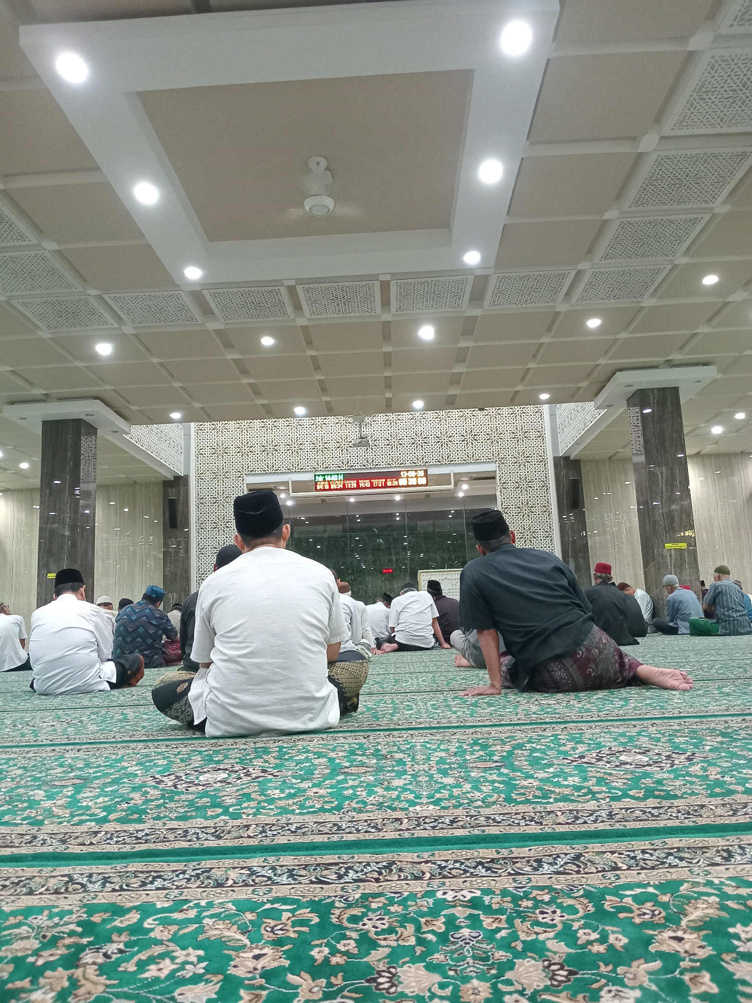 A low-angle, full shot captures the interior of a mosque with men seated on a green carpet for prayer. In the foreground, a man in a white shirt and black cap faces away from the camera, his legs crossed on the carpet. To his right, another man in a black shirt and patterned sarong sits with his legs extended. The green carpet features a floral pattern with beige and darker green accents.
In the middle ground, various men are seen in prayer positions, some wearing white shirts and caps, others in traditional clothing. The mosque's architecture is evident with tall, patterned columns on either side of a large decorative screen with geometric designs. A digital display board with red lettering is visible in the background, indicating prayer times.
The ceiling is a prominent feature, with a modern design incorporating square panels, some featuring intricate white geometric patterns, and recessed lighting. A ceiling fan is also visible. The environment appears to be a well-lit, contemporary mosque hall. The city of Randuagung, Indonesia is not directly depicted or identifiable from the visual elements within the image.