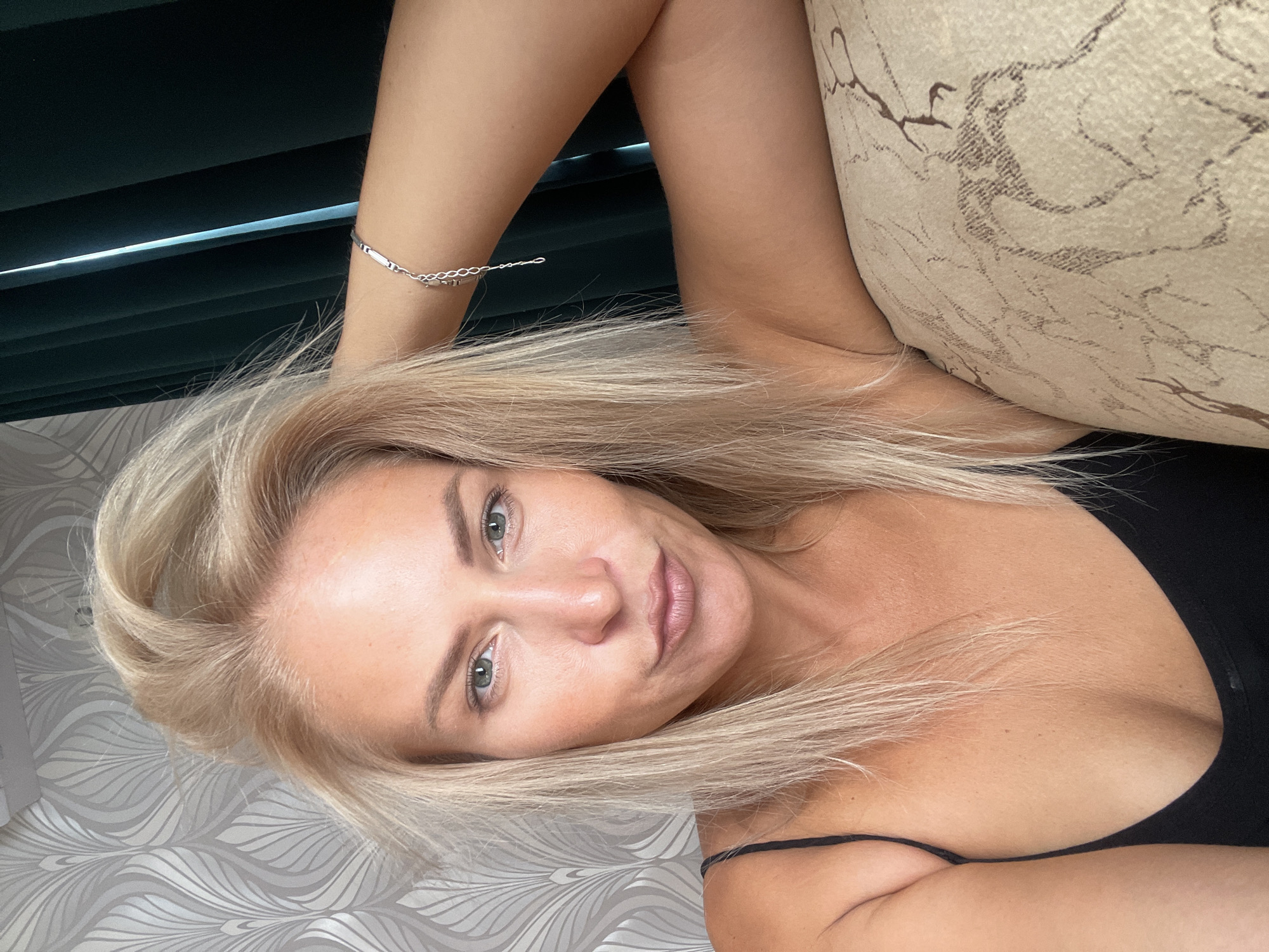 A blonde woman is captured in a close-up, selfie-style shot, leaning back on a couch with her arm behind her head. She is wearing a black camisole. Her hair is styled loosely around her face. The background features a patterned wallpaper and dark green curtains. The image does not provide enough information to determine the location in Krasnodar, Russia.