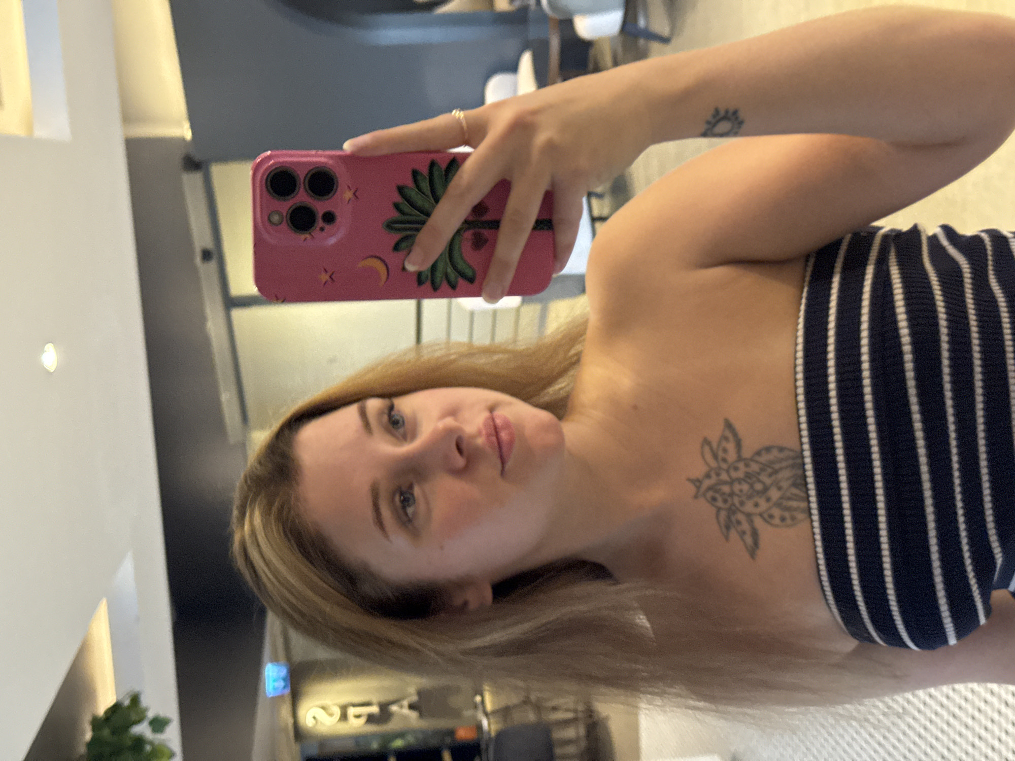 A blonde woman with blue eyes takes a mirror selfie in a well-lit indoor space. She is wearing a strapless, navy and white striped top and has a tattoo on her chest of an abstract, floral design. She is also wearing a ring on her right ring finger and has a small sun tattoo on her left forearm. She holds a pink phone with a palm tree and celestial design in her right hand. In the background, there are hints of a modern interior with reflective surfaces and ambient lighting. The location is identified as Aksu, Turkey.