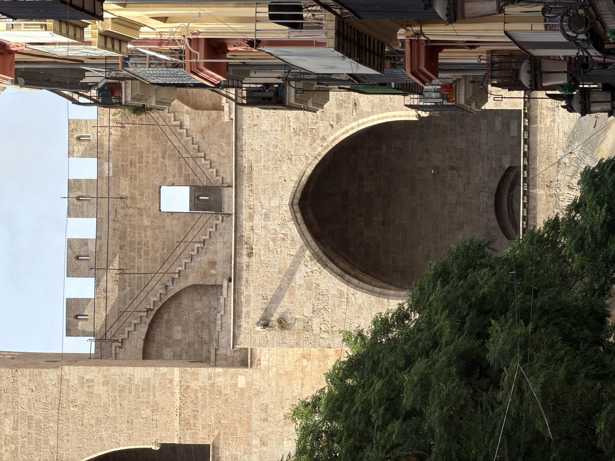 A view of the Serrans Towers in Valencia, Spain, is captured from a street level perspective. The imposing stone structure of the towers dominates the left side of the frame, featuring a prominent arched entrance and an external staircase leading to battlements. To the right, a narrow street is lined with colorful buildings exhibiting ornate balconies and architectural details, including lamps and flower pots. Lush green trees partially obscure the lower portion of the towers and the street. The scene is bathed in natural daylight, casting shadows on the stone facade and highlighting the textures of the architecture and foliage. The sky is clear with a pale, overcast appearance. No people are visible in the image.