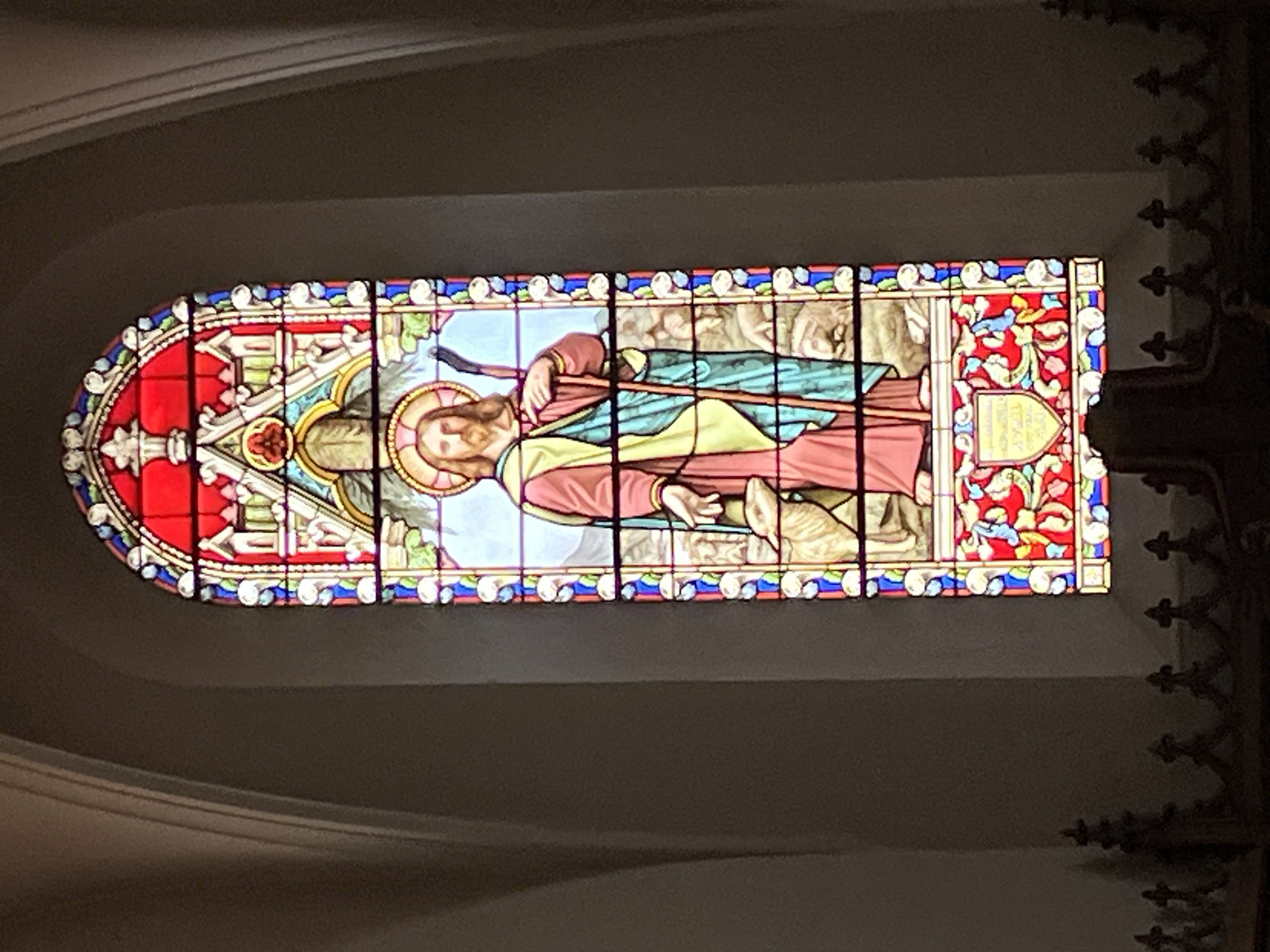 A stained glass window depicts Jesus as the Good Shepherd, holding a crook and with a sheep at his feet. He has a halo, and other sheep are in the background. The window is set within the architecture of a church, located in Chambœuf, France. The surroundings are dark, but the glass illuminates the interior.