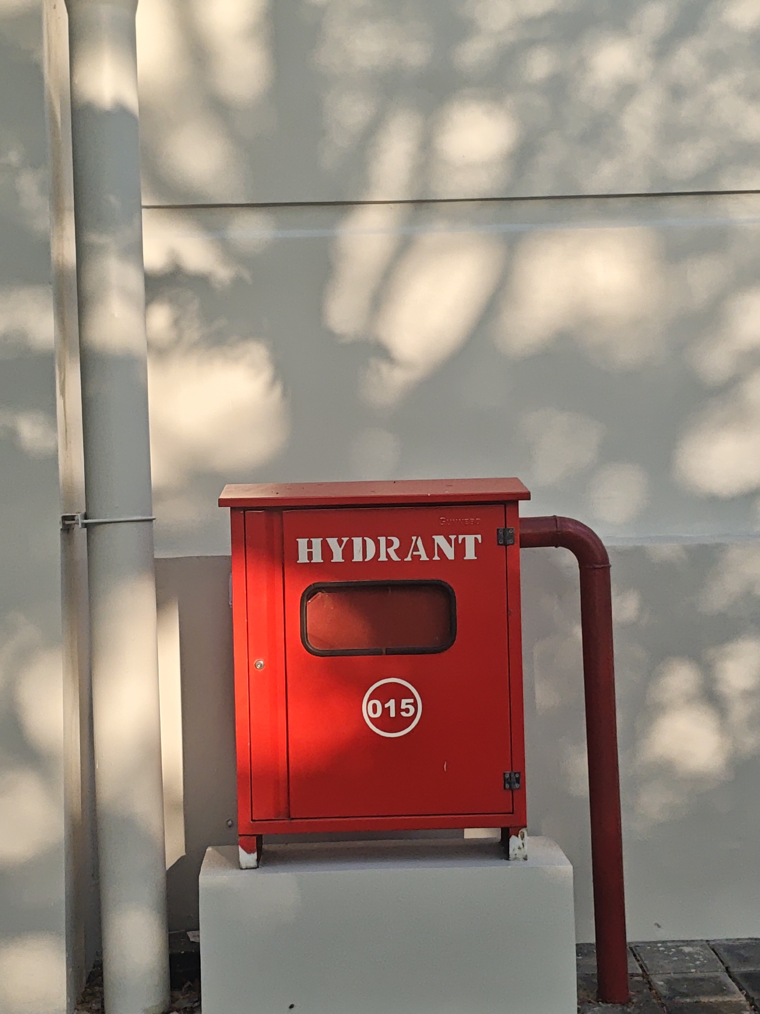 The image shows a bright red fire hydrant box with the word "HYDRANT" printed in white capital letters above a rectangular window. A white circular emblem with the number "015" is affixed to the front of the box. The box is mounted on a white concrete pedestal. To the left of the fire hydrant box, a large white pipe runs vertically, attached to a wall by a metal bracket. To the right, a red pipe emerges from the side of the fire hydrant box and curves downwards. The background is a light-colored wall with dappled shadows cast by sunlight and foliage. The ground beneath the hydrant is paved with dark rectangular tiles, and there is some debris. The scene appears to be outdoors in a well-lit area, possibly on a street or public space. There are no people visible in the image. The location is identified as Bae, Indonesia.