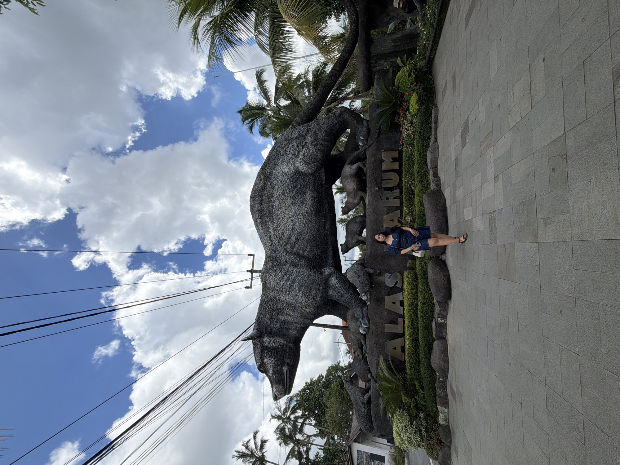 A woman stands to the right of a grey stone sculpture in Tegal Suci, Indonesia. The sculpture, featuring a large bear-like animal and other smaller figures, is positioned next to a sign bearing the text "ALAS RARUM." The area is paved with grey stone tiles and lined with greenery. Above, palm trees and electrical wires are visible against a backdrop of blue sky and white clouds.