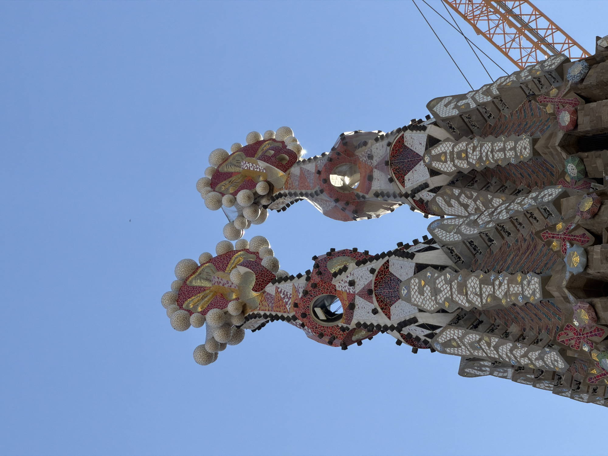 The image shows the Sagrada Familia in Barcelona, Spain, specifically focusing on two of its distinctive spires. These spires are intricately decorated with mosaic tiles and spherical elements, resembling stylized fruits or flowers at their apex. The architecture is elaborate and colorful, with geometric patterns and contrasting textures. A bright blue sky serves as the backdrop, with a few clouds visible. To the right of the spires, a section of an orange construction crane is visible, suggesting ongoing work or restoration at the basilica. The perspective is from a low angle, looking upwards at the towering structures.