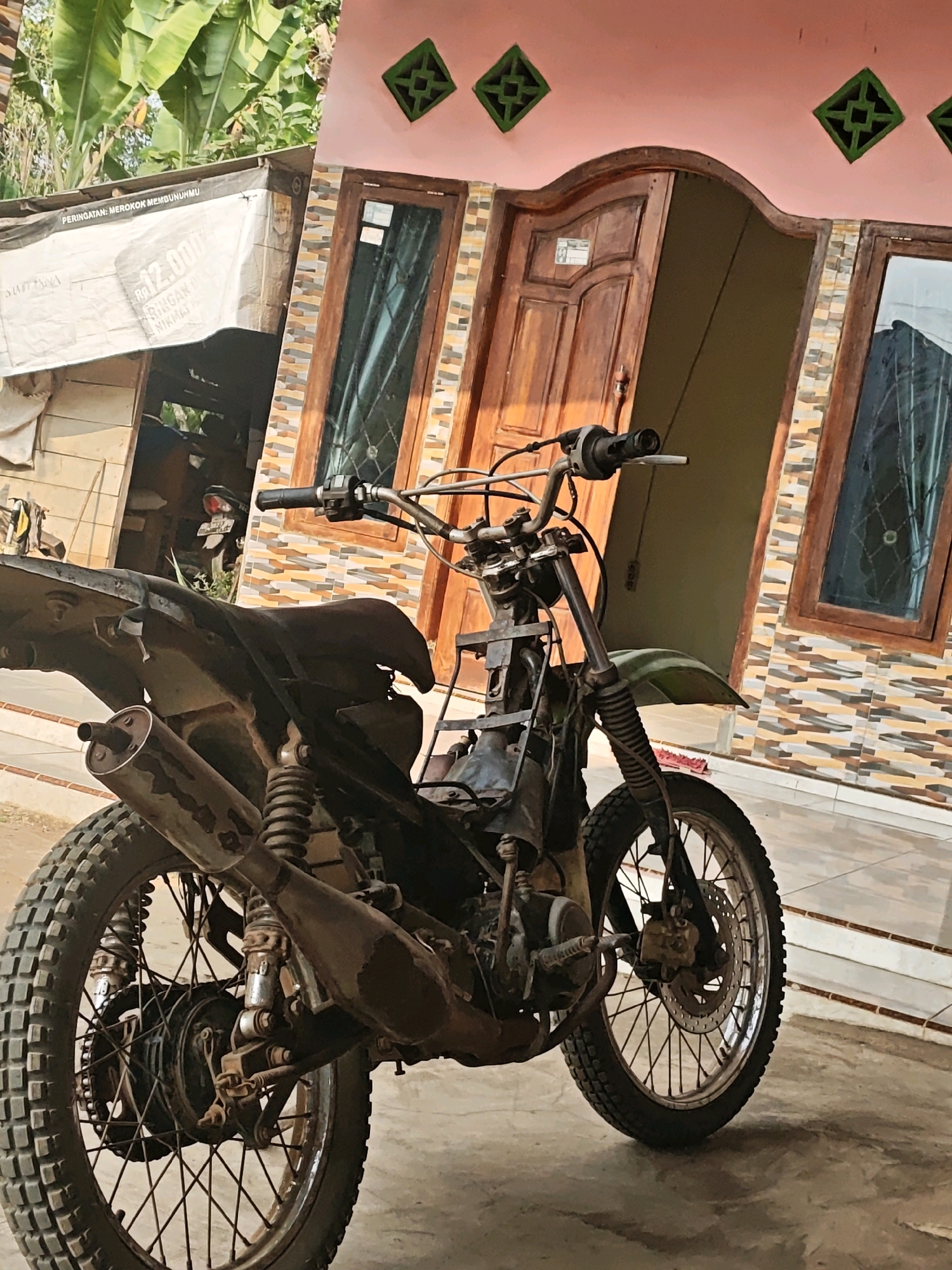 A slightly tilted, medium shot captures a weathered, black dirt bike parked on a concrete surface in front of a house. The house features a light pink facade adorned with decorative green diamond-shaped vents and a brown wooden door with a curved top, flanked by a wooden-framed window. The lower portion of the house is tiled with horizontal bands of beige, brown, and grey. To the left of the house, a makeshift structure covered with a white tarp, bearing Indonesian text and a monetary figure, is partially visible. Large green banana leaves are prominent in the upper left background. The dirt bike, showing signs of wear and tear, has a black seat, a protruding exhaust pipe, and knobby tires. The handlebars and front suspension are visible. The scene appears to be an outdoor setting in Karang Sari, Indonesia, likely a residential area.