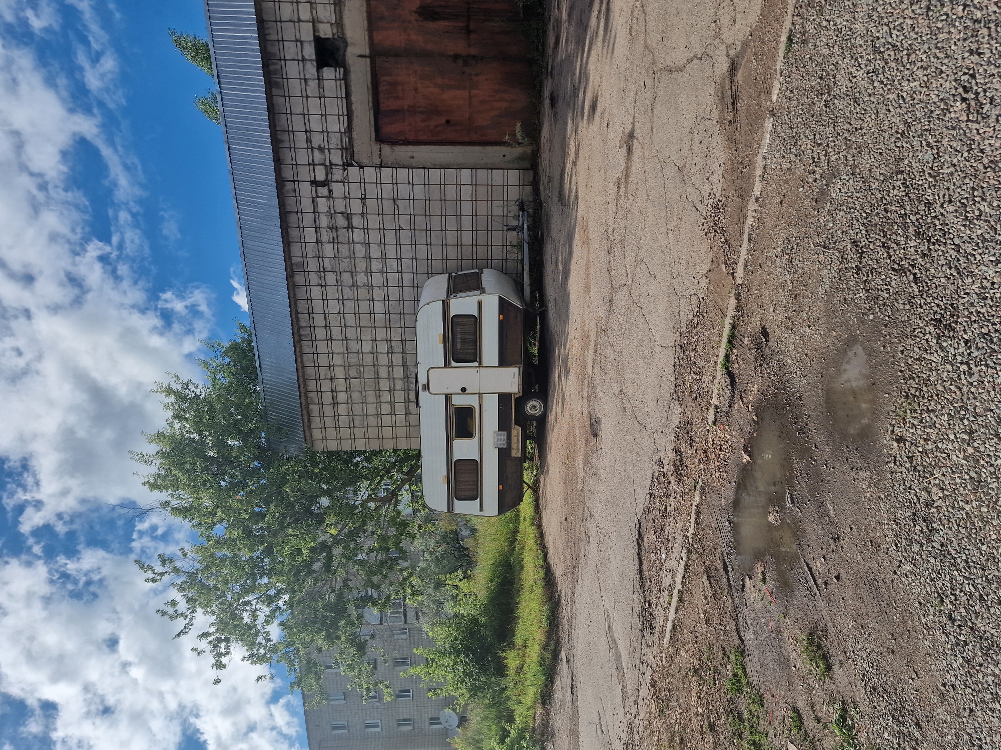 The image shows a small white caravan parked next to a building with a tiled facade. The building has a metal roof and a large, reddish-brown garage door. A tree with lush green leaves stands between the caravan and a multi-story apartment building in the background. The sky is blue with white clouds.
The caravan is white with brown trim and has several windows. It is hitched to a trailer. The ground in front of the caravan is paved with asphalt and gravel. There is a puddle of water on the asphalt. The scene appears to be outdoors, likely in a residential or industrial area.
There is no visible text in the image. The lighting suggests it is daytime. The weather appears to be clear and sunny. The overall impression is one of a quiet, possibly rural or suburban, setting.