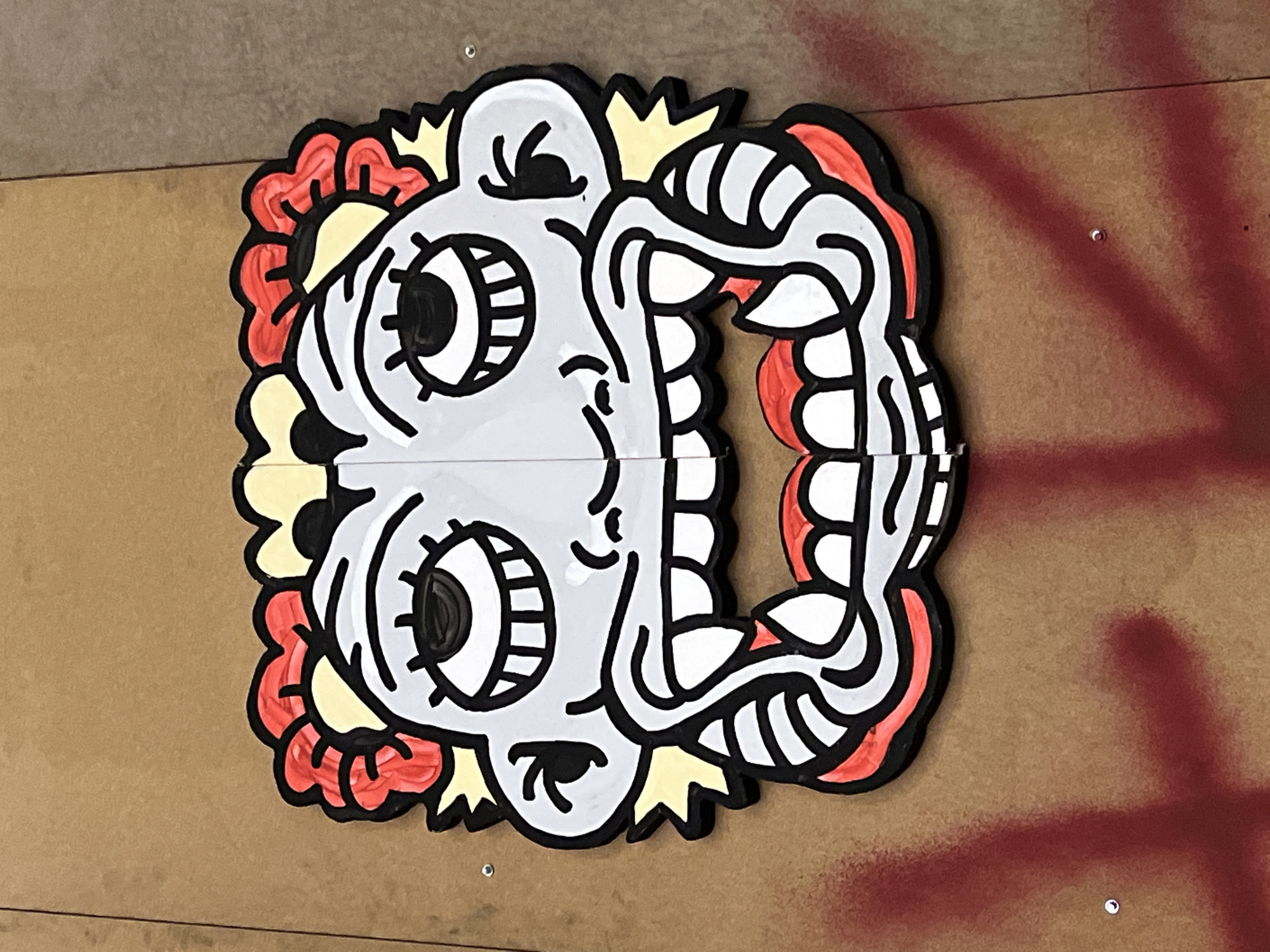 The image is a close-up shot of a cartoonish graffiti mural on a wooden panel. The mural depicts a grey face divided horizontally, seemingly split into two separate panels. The face has large, round eyes with multiple concentric circles for detail, and a wide, open mouth exposing numerous square teeth. The mural is outlined in black and embellished with red and yellow floral or flame-like designs around the perimeter of the face. The wooden panel it is painted on is a light brown color and has some spots of red paint spray near the right edge of the image. The mural appears to be mounted on the wood with visible screws near the lower-left and upper-left edges. Based on the location, it could be placed anywhere in Madrid, Spain.