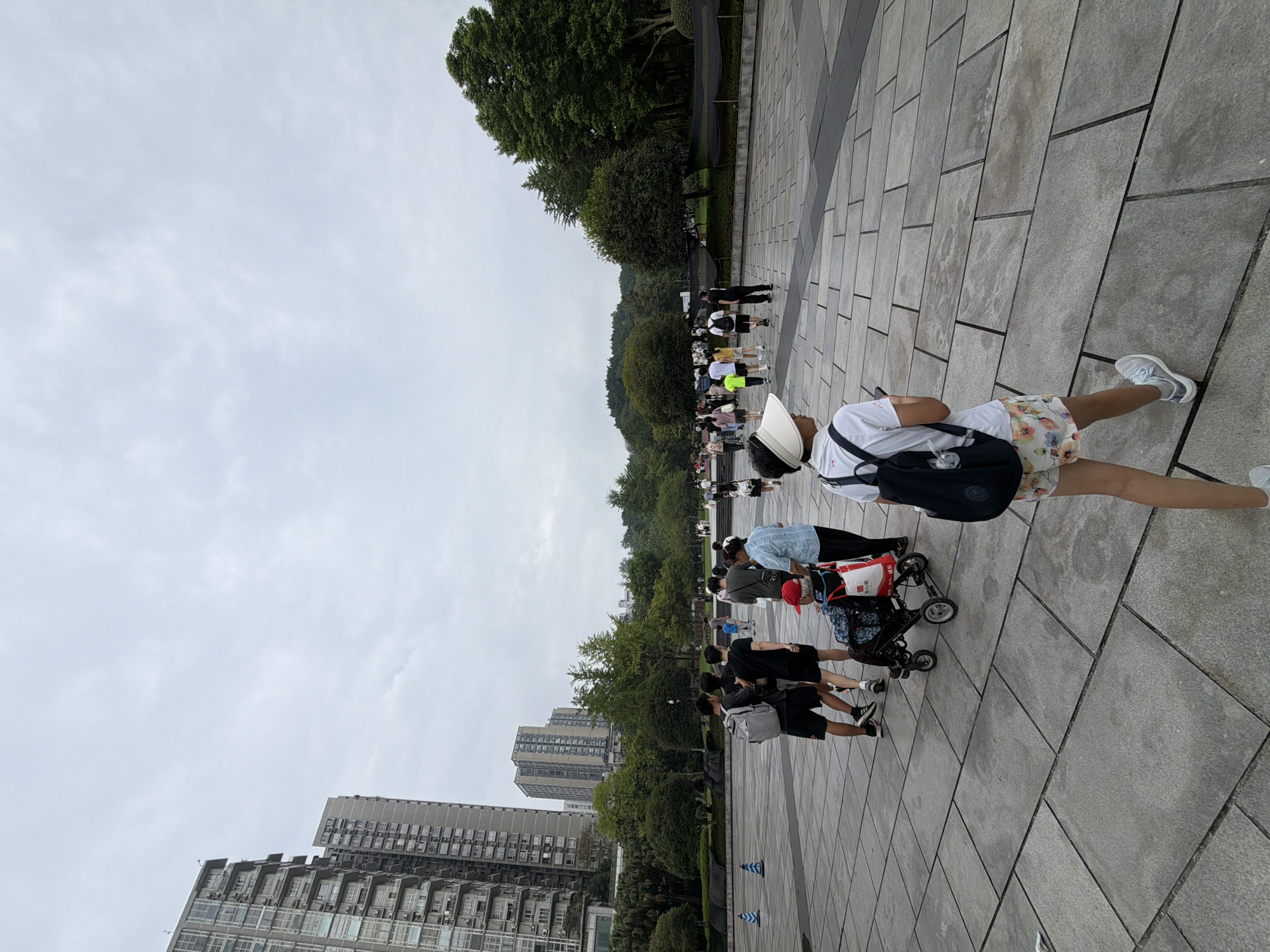 In the Jiulongpo District, China, an outdoor scene unfolds under an overcast sky, with a wide expanse of grey paving stones dominating the foreground. Several individuals are present, engaged in various activities. In the mid-ground, a group of three young men walks away from the viewer; the one on the left carries a grey backpack. To their right, a woman pushes a stroller containing a child, and an elderly person in a wheelchair, wearing a red cap and holding a white bag with red lettering, is positioned nearby. Further into the scene, a procession of people walks towards a park-like area with numerous trees and undulating terrain in the background. In the far distance, several tall, modern apartment buildings rise above the treeline, indicative of an urban environment. The perspective is from ground level, looking slightly upwards towards the sky and the buildings.