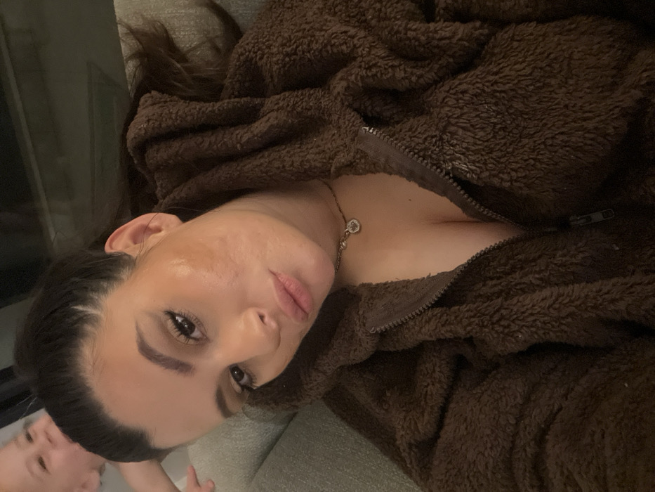 An indoor scene features a female subject positioned centrally, capturing a selfie from a slightly elevated angle. She has dark hair pulled back and wears a dark brown, textured, hooded jacket, partially unzipped to expose her upper chest and a silver-toned chain necklace with a small, circular, possibly jewel-set pendant. Her face exhibits cosmetic enhancements, including defined eyebrows, winged eyeliner, and lipstick. A pale, irregularly shaped mark is visible on her right cheek, alongside minor skin textural variations. Her gaze is directed towards the camera, with a slight pout. In the upper left background, a baby or young child with fair skin is partially visible, appearing to smile and look towards the left. The immediate environment includes blurred light-colored upholstery on the left and dark, indistinct background elements. The image provides no discernible visual information to specify a location within Kiryat Bialik, Israel.