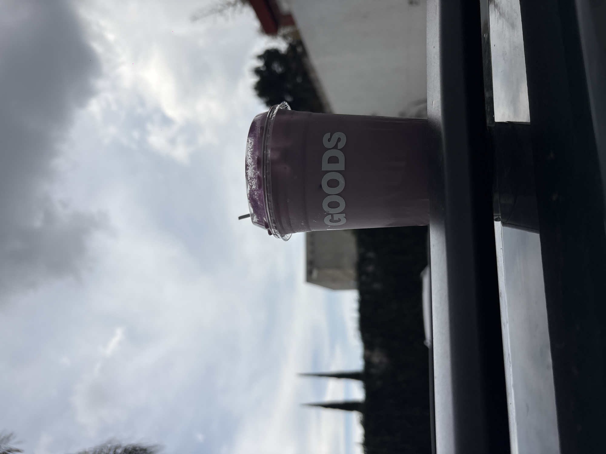 A plastic cup with the word "GOODS" printed on its side sits on a ledge. The cup contains a purple-colored beverage, possibly a smoothie. The background shows an overcast sky with trees and a building. The location is likely Puebla, Mexico.