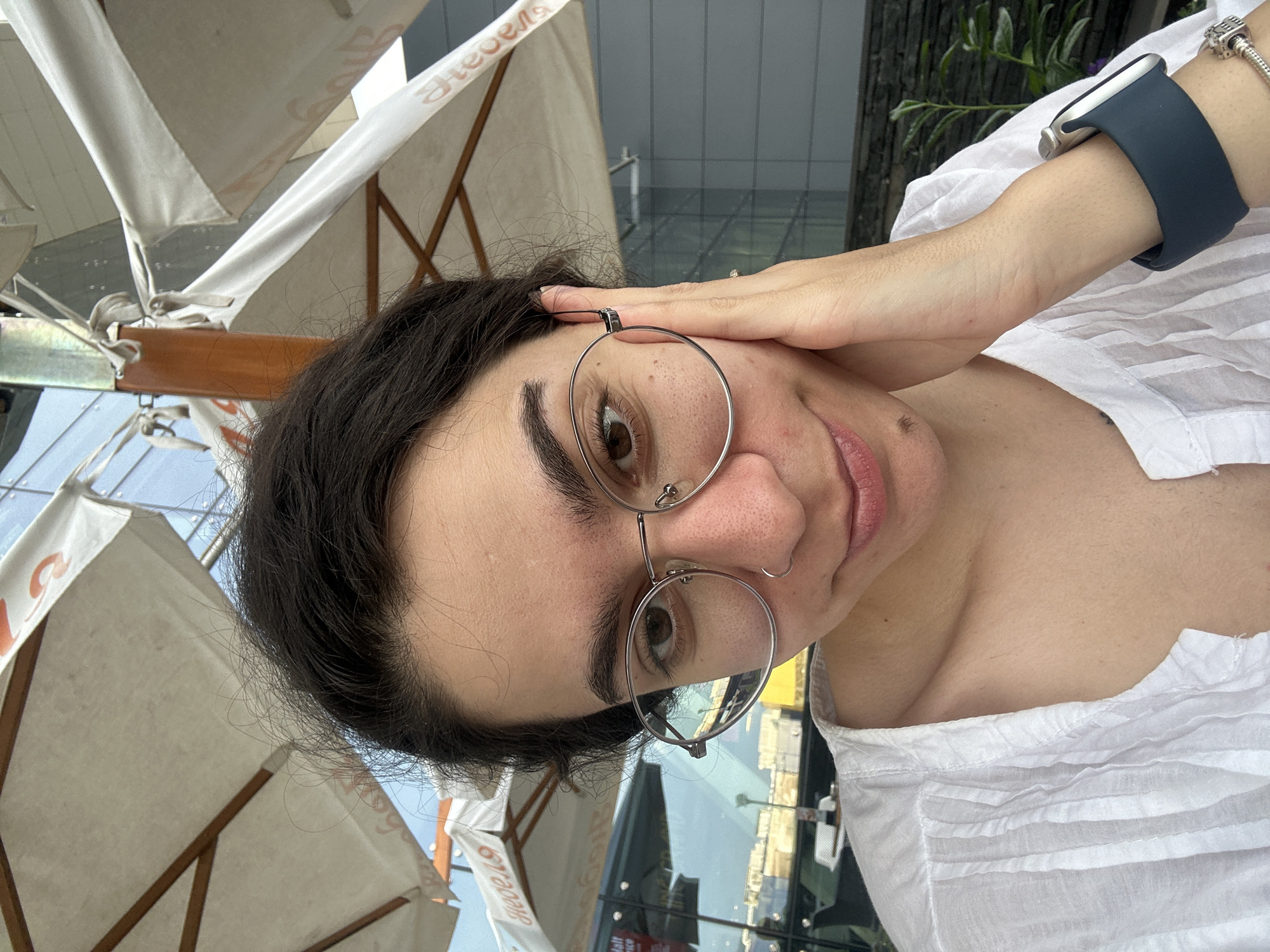This is a selfie taken outdoors, likely at a cafe, with large white umbrellas providing shade. The subject is a young woman with dark hair, wearing large, round, silver-rimmed glasses and a white, crinkled-fabric shirt. Her right hand is resting on her cheek. She is wearing a dark blue watch on her left wrist and a charm bracelet on her right wrist. The background shows buildings with glass facades and some greenery. The text visible on the umbrellas appears to be in Cyrillic, suggesting the location is in Sofia, Bulgaria. The overall lighting indicates it is daytime.