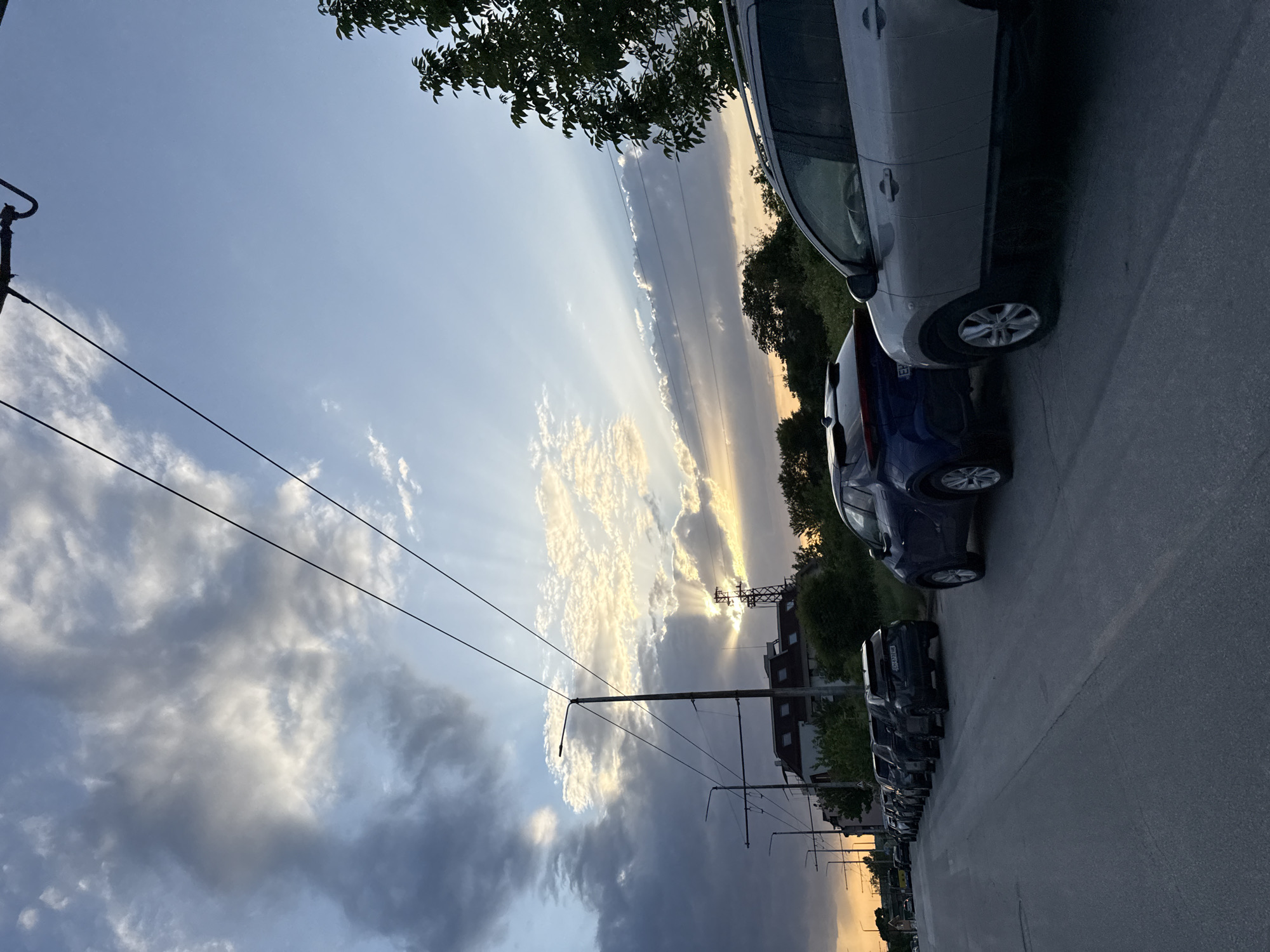 The image shows a street in Pleven, Bulgaria, at sunset. The sky is filled with clouds and sun rays breaking through. On the right side of the image, cars are parked along the street. Power lines are visible in the sky. Buildings are seen behind the parked cars. The street appears to be paved and well-maintained.
