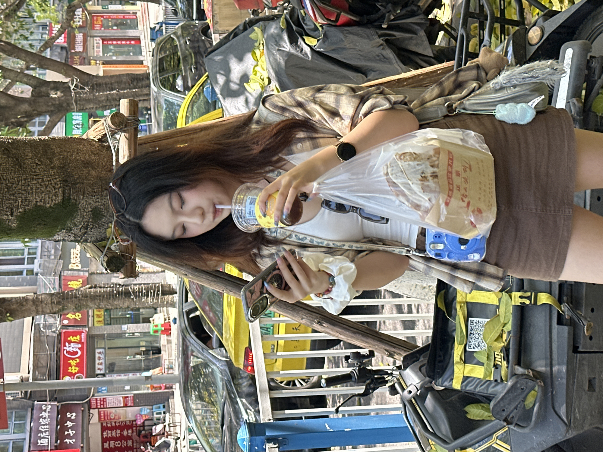 A young woman with long brown hair, wearing a white top, a brown skirt, and a plaid shirt, sits on a bench next to a tree. She is drinking from a clear plastic cup with a straw while holding a smartphone. A plastic bag containing food sits on her lap, partially obscuring her outfit. The scene is set outdoors in the Shapingba District of China, as evidenced by the Chinese signage in the background. The background includes various shop signs in Chinese characters and several parked vehicles, including a yellow taxi. A scooter is visible near the bench. The light suggests it is daytime.