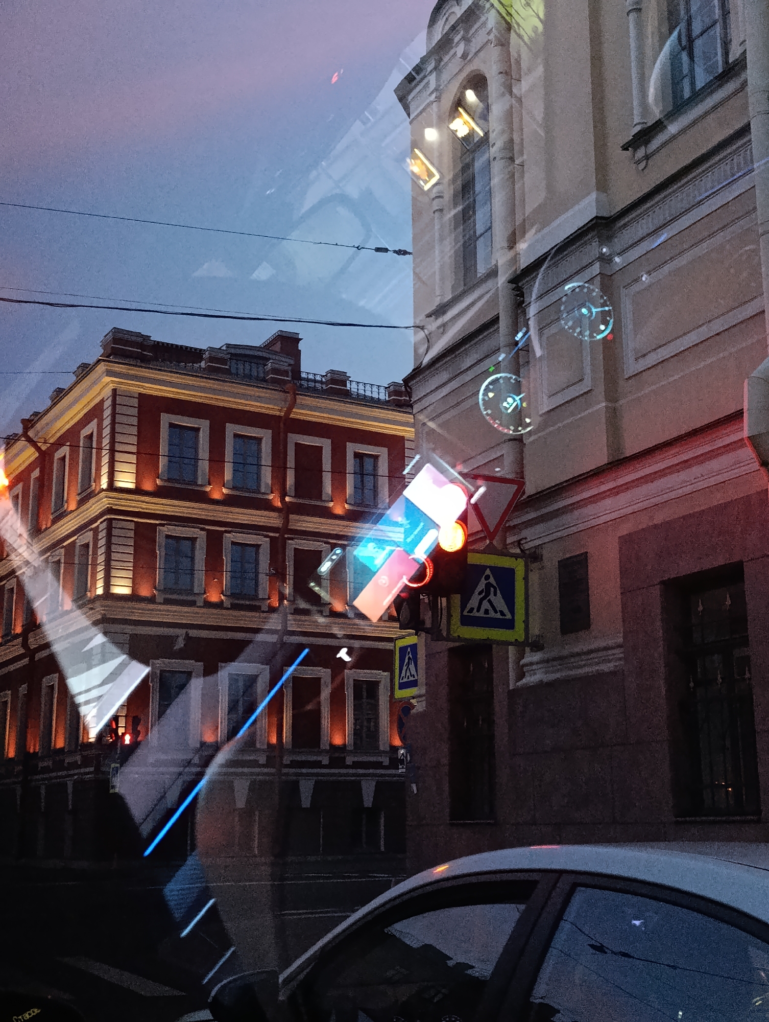 This is a reflective image of a street scene in Saint Petersburg, Russia, taken from inside a vehicle. The primary focus is on the architecture and traffic signals of the city. On the left, a multi-story red building with illuminated windows and decorative white trim is visible. To the right, a light-colored building with arched windows and classical architectural elements dominates the background.
Overlaying the scene are reflections, most prominently a bright blue diagonal line and geometric light patterns. Traffic signals, including a pedestrian crossing light showing red and yellow, and a triangular warning sign, are also present. A pedestrian crossing sign with a yellow background and a silhouette of a person walking is visible.
Digital overlays appear on the buildings, resembling futuristic interfaces with circular gauges and glowing elements, suggesting this might be a digitally augmented reality capture. The sky is a dusky blue with hints of pink, indicating it is either early morning or late evening. A portion of a silver car's interior and exterior is visible in the foreground. The overall impression is a blend of historical urban architecture and modern digital aesthetics.