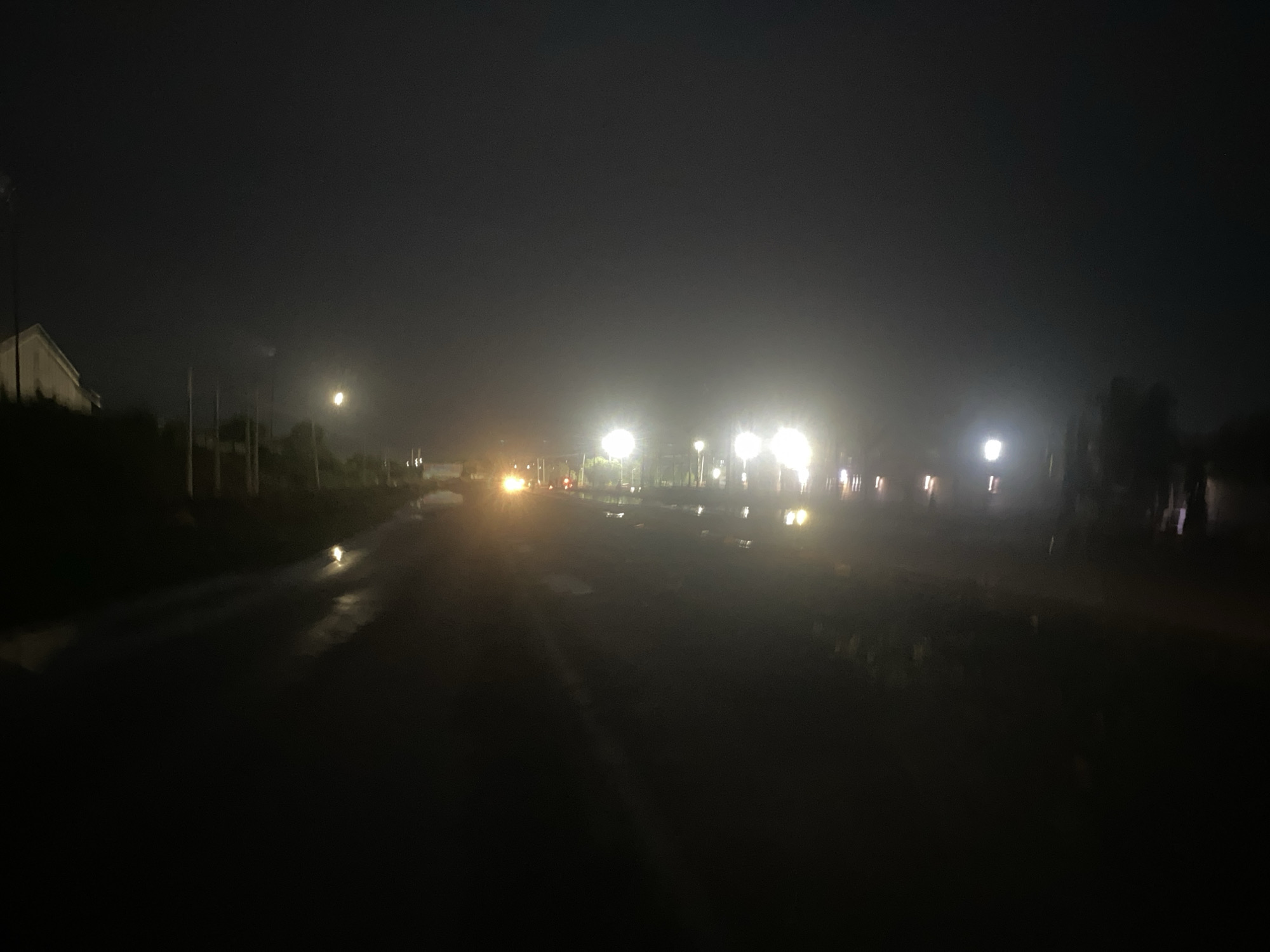A dark, overcast night scene in Karmajiji, Nigeria, focuses on a wet, paved road stretching into the distance. Streetlights line the road, their illumination blurring due to atmospheric conditions. Vehicle headlights are visible further down the road. On the left side of the road, a building is faintly discernible. The road surface reflects the sparse light sources. The foreground is predominantly dark, obscuring details.