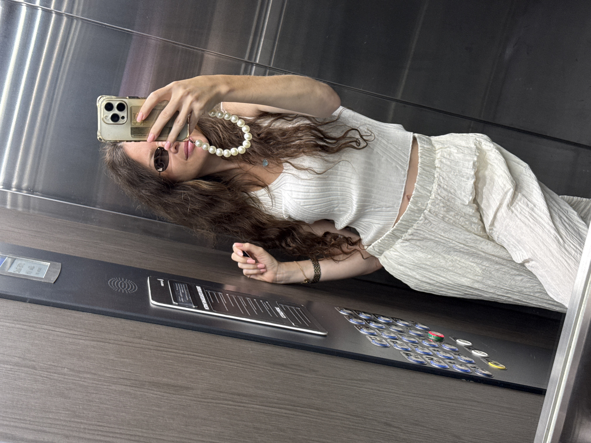 In an elevator in the city of Khimki, Russia, a woman with long, wavy brown hair takes a selfie. She is wearing a white ribbed tank top, white pleated pants, and small oval sunglasses. Around her neck, she wears a strand of large pearls. She is holding a smartphone in her right hand, which has a gold case and multiple camera lenses. The elevator walls are made of brushed stainless steel and wood paneling. On the left side of the elevator, there is a control panel with numerous buttons. The woman's left hand rests on the panel, and she is wearing a bracelet on her wrist.