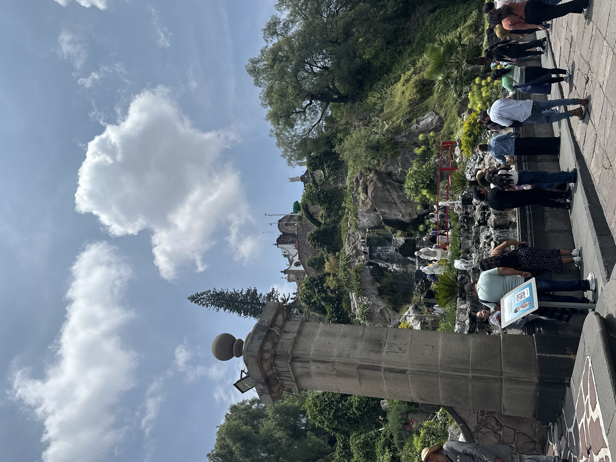 This is a medium-shot photograph of the Basílica de Santa María de Guadalupe in Mexico City, Mexico. The scene captures a portion of the exterior grounds, with a focus on a man-made waterfall cascading down a rocky hillside. Several people are visible, some standing near the waterfall and others walking along a paved pathway in the foreground. In the foreground, a large stone pillar with a spherical finial stands prominently on the left. To the right of the pillar, a family is gathered around an information kiosk displaying a QR code and an image of a child. The hillside is covered in lush green vegetation, with trees and various plants growing on the slopes. At the top of the hill, a white building with architectural details that suggest a church or basilica is visible. The sky above is partly cloudy with large, white cumulus clouds against a bright blue backdrop. The overall atmosphere appears to be that of a sunny day at a popular religious or tourist site.