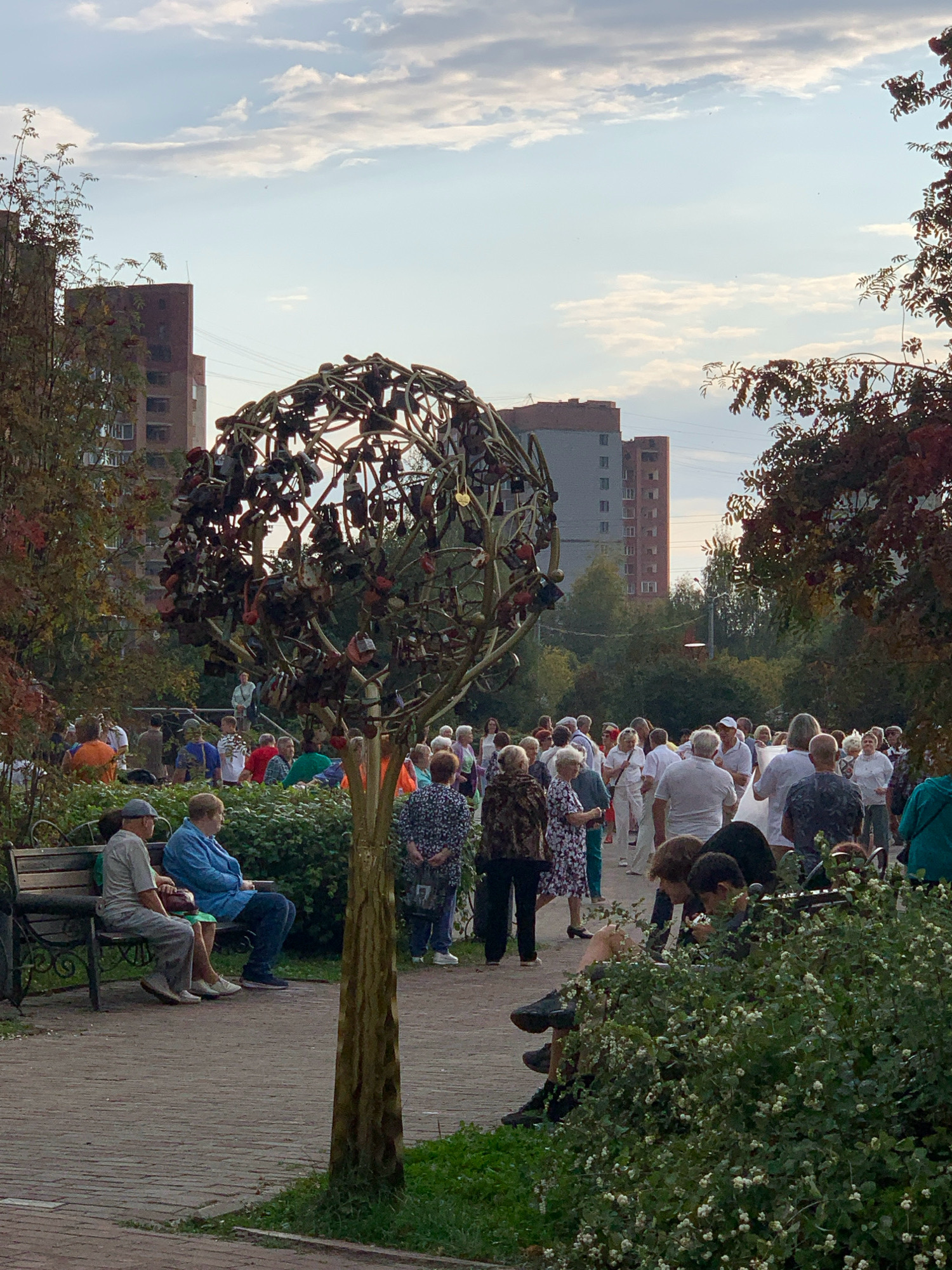 The image captures a public park scene in Prokopyevsk, Russia, featuring a large, spherical metal sculpture adorned with numerous padlocks. A diverse group of people, predominantly adults, are dispersed throughout the park, some standing in clusters and others seated. In the foreground, two individuals are seated on a park bench, facing away from the camera. Behind them, a crowd mingles near the padlock sculpture, with others visible in the background. The environment consists of a paved walkway, lush green bushes, trees with autumnal foliage, and tall residential buildings. The sky above is partly cloudy, suggesting it is daytime.