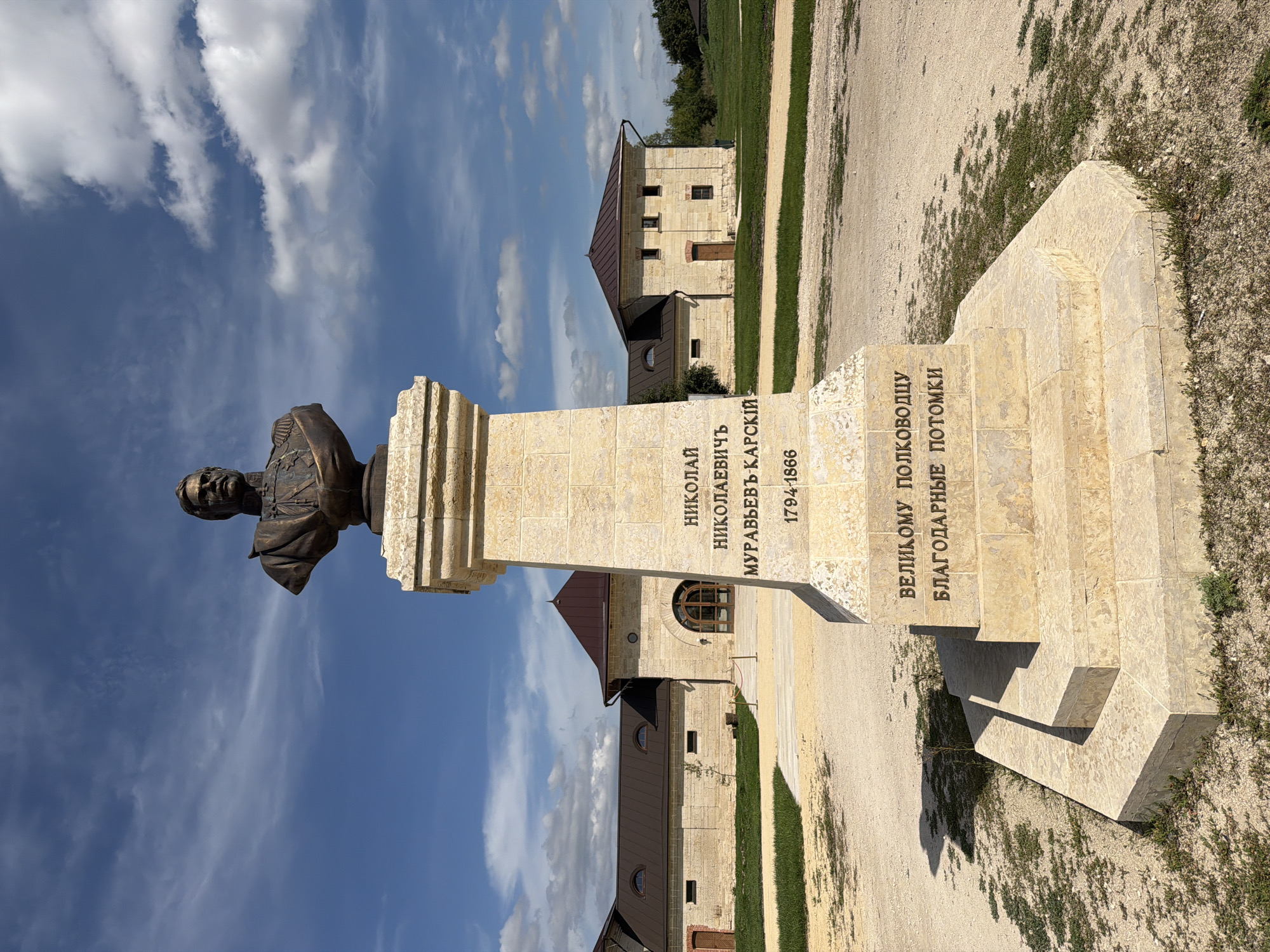 A bronze bust of a man with a mustache and wearing a military uniform is mounted on a stone pedestal in an outdoor setting. The pedestal is inscribed with Russian text and dates, "НИКОЛАЙ НИКОЛАЕВИЧ МУРАВЬЕВ-КАРСКИЙ 1794-1866" and "ВЕЛИКОМУ ПОЛКОВОДЦУ БЛАГОДАРНЫЕ ПОТОМКИ". The bust and pedestal are situated on a gravel path with some sparse vegetation. In the background, there are traditional Russian-style stone buildings with brown roofs and arched doorways, surrounded by green grass and trees under a blue sky with scattered white clouds. The location is identified as the village of Skornyakovo, Russia.
