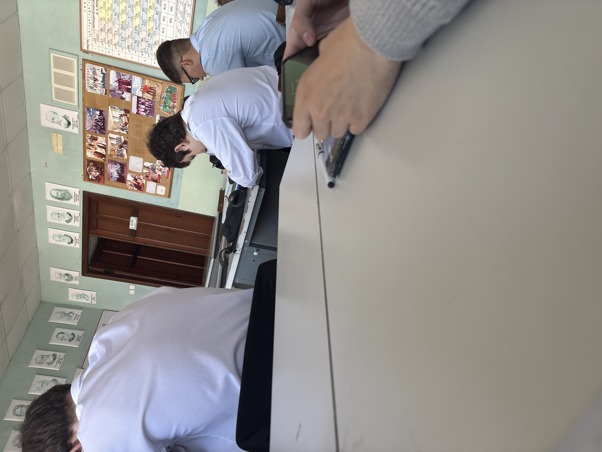 The image captures a classroom or laboratory scene, viewed from a close perspective over a desk. In the immediate foreground, a person's hands are visible on a light-colored desk, holding a black smartphone. A black marker lies on the desk beside the phone. To the right, the back of a student's head and shoulders are visible; this student is wearing a white shirt and leaning forward over a desk.
Further into the room, two other male students are present. One, wearing a light blue shirt and glasses, is also leaning over a desk, looking down. Another student in a white shirt is positioned slightly in front of him, similarly focused on something on a desk.
The walls of the room are a light green or teal color. A large Periodic Table of Elements is displayed on the back wall, partially labeled "ПЕРИОДИЧЕСКАЯ СИСТЕМА Д. И. МЕНДЕЛЕЕВА" (Periodic System of D. I. Mendeleev). Below this chart, a cork bulletin board is filled with numerous photographs, suggesting class activities or events. To the left of the bulletin board, several framed monochrome portraits of historical figures or scientists adorn the wall; one portrait clearly shows the dates "1829-1896". A wooden door and a ventilation grate are also visible on the wall.
The bright lighting suggests it is daytime. The students appear to be focused on a task, possibly engaged in a lesson or an experiment.
Visible text includes:
- On the periodic table: "ГРУ", "ПЕРИОДИЧЕСКАЯ СИ", "Д. И. МЕНД", chemical symbols (e.g., Na, Mg, Al, Si, K, Ca, Sc, Ti, Cu, Zn, Ga, Ge, Rb, Sr, Y, Zr, Ag, Cd, In, Sn, Sb, Au, Hg, Tl, Pb), formula examples (R, O, R₂O, RO, R₂O₃, RO₂, RH, RH₃, RH₄), "ЛАНТАНОИДЫ", and "АКТИНОИДЫ".
- Below a portrait: "1829-1896".