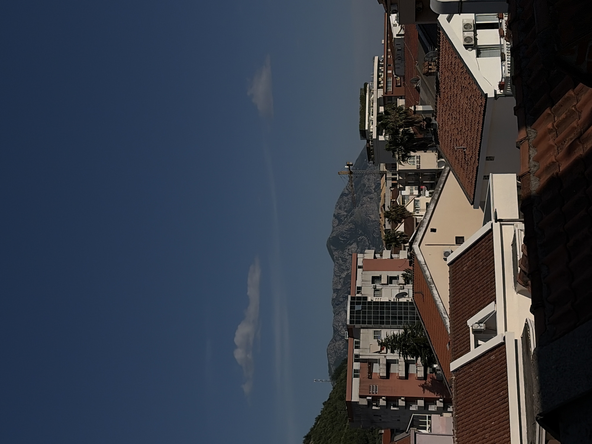 The image shows a view over the city of Budva, Montenegro, with residential buildings and terracotta-tiled roofs dominating the foreground and midground. Behind the buildings, a mountain range rises, and a clear blue sky with some scattered clouds can be seen. A construction crane is visible amidst the buildings.